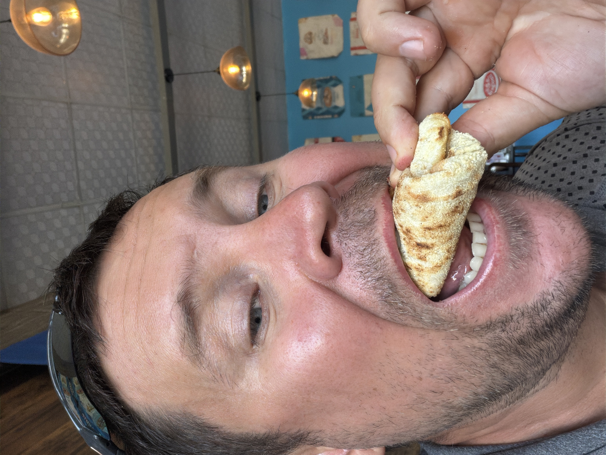 A man, seen from the chest up, is taking a bite of a grilled flatbread in what appears to be a restaurant. He is looking into the camera with his mouth open and the flatbread partially in his mouth. His right hand is holding the flatbread to his mouth. He has short brown hair, stubble on his chin, and is wearing a grey patterned shirt. In the background, there are light fixtures and a turquoise wall with some decorations. The overall lighting is warm. The scene is likely indoors.