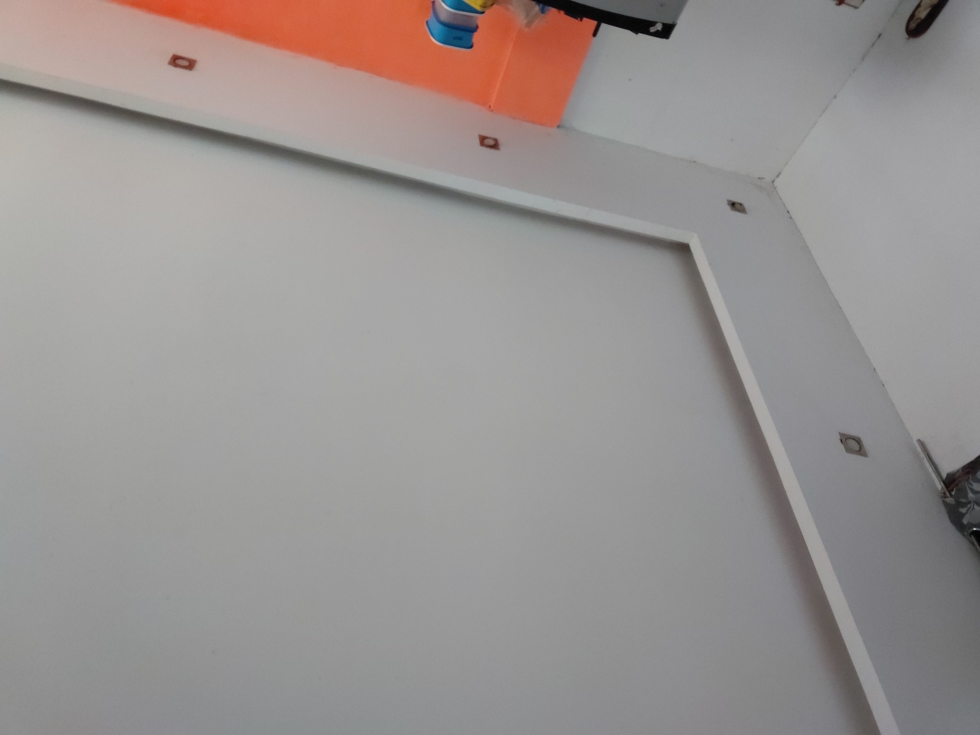 The image captures an interior view, likely a room or hallway, characterized by predominantly light-colored walls and ceiling. The perspective is upward-looking, into a corner of the space.
The main wall on the left and center is a plain light grey or off-white. A distinctive white, recessed frame or molding defines a large rectangular area on this wall. Along the top edge of this frame and continuing onto the ceiling, as well as along the right side of the frame, are several small, square-shaped cutouts, which appear to be housings for recessed lighting fixtures, some with a dark interior visible.
Above the white frame, in the upper-left portion of the image, is a section of wall painted a vibrant orange. Suspended near the junction of this orange section and the ceiling is a small object with blue and yellow components, possibly part of a tool, container, or toy.
The ceiling is white. In the upper-right corner, where the white ceiling meets the right wall, some indistinct dark objects are visible, which could be wires, equipment, or debris. One crop reveals a round, metallic, possibly grimy object, which might be an old light fixture or vent opening. The wall on the far right is also a light grey or off-white.
No activity or people are present. The lighting suggests daytime or bright artificial illumination. There is no visible text in the image. The exposed elements and cutouts might indicate that the space is under construction, renovation, or is a utility area.
