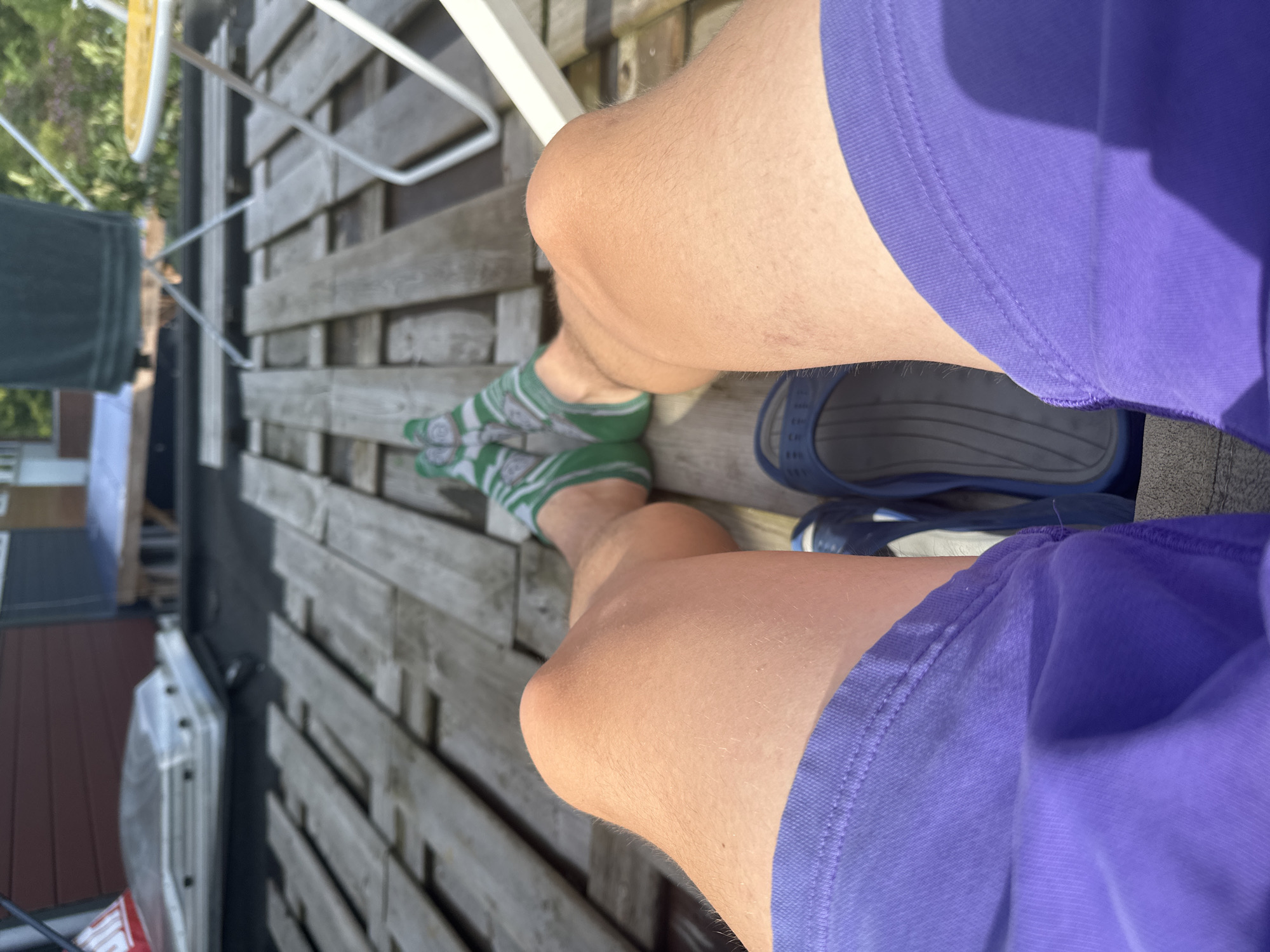A first-person perspective shot shows a person's legs and feet resting on a wooden deck, possibly a balcony or rooftop, in Lelystad, Netherlands. The person is wearing purple shorts and green socks with a white pattern. A blue sandal is visible on the right foot. The background suggests an outdoor setting with glimpses of a building, greenery, and outdoor furniture, indicating a residential or recreational area in the city. The lighting suggests it is daytime, possibly late morning or afternoon.