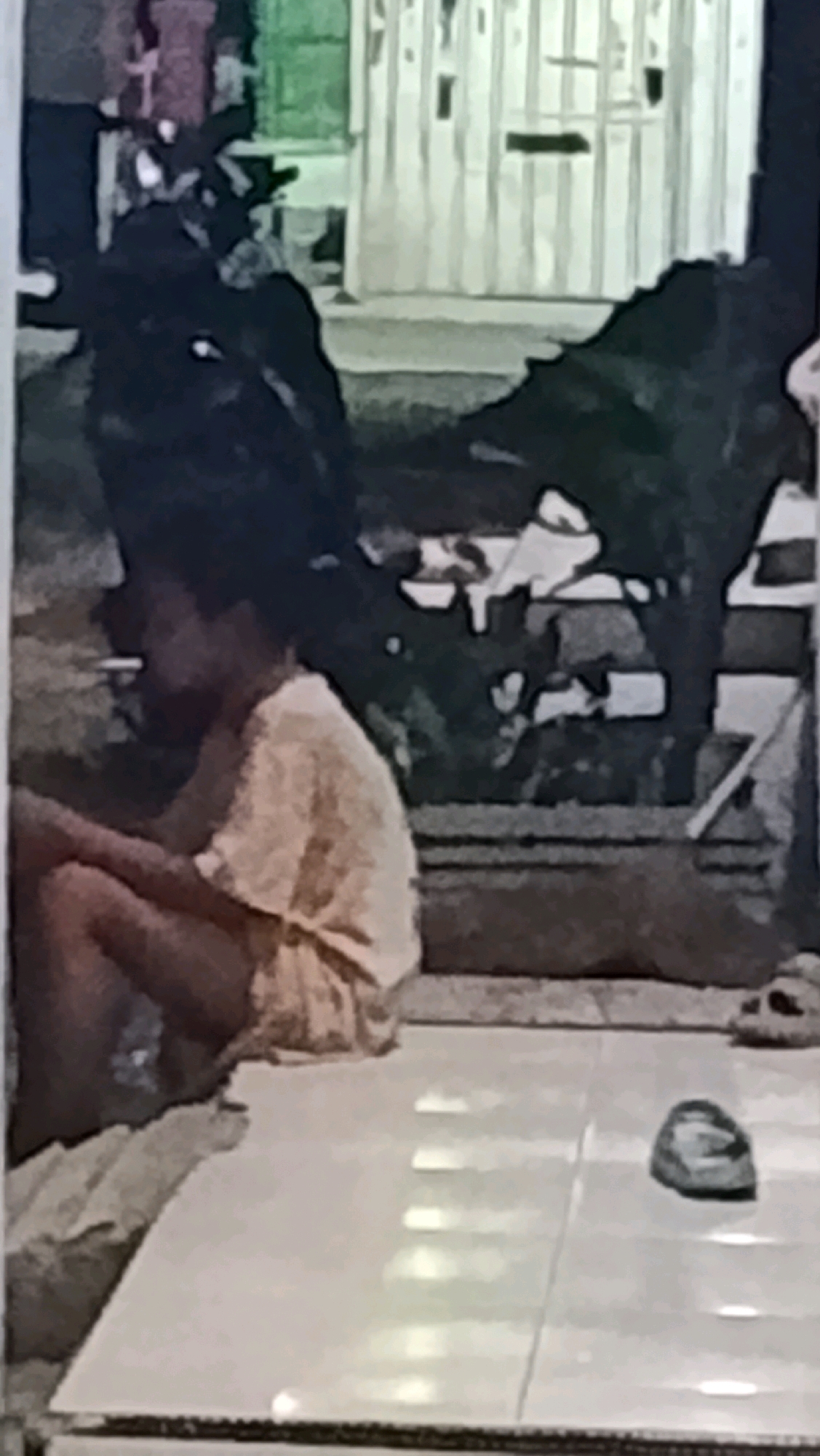 A person is sitting on a tiled floor in what appears to be an outdoor or semi-outdoor setting. They are wearing a light-colored, possibly beige, t-shirt and dark shorts. The person is holding a cigarette in their mouth and appears to be smoking. Their legs are bent, and they are hunched forward. In the background, there are dark, indistinct shapes that could be furniture or other objects, and a building with vertical lines, possibly a corrugated metal wall or a fence. There is also a shoe visible on the tiled floor to the right. The overall lighting suggests it is either dusk or a dimly lit area. The location is identified as Ujungrusi, Indonesia.