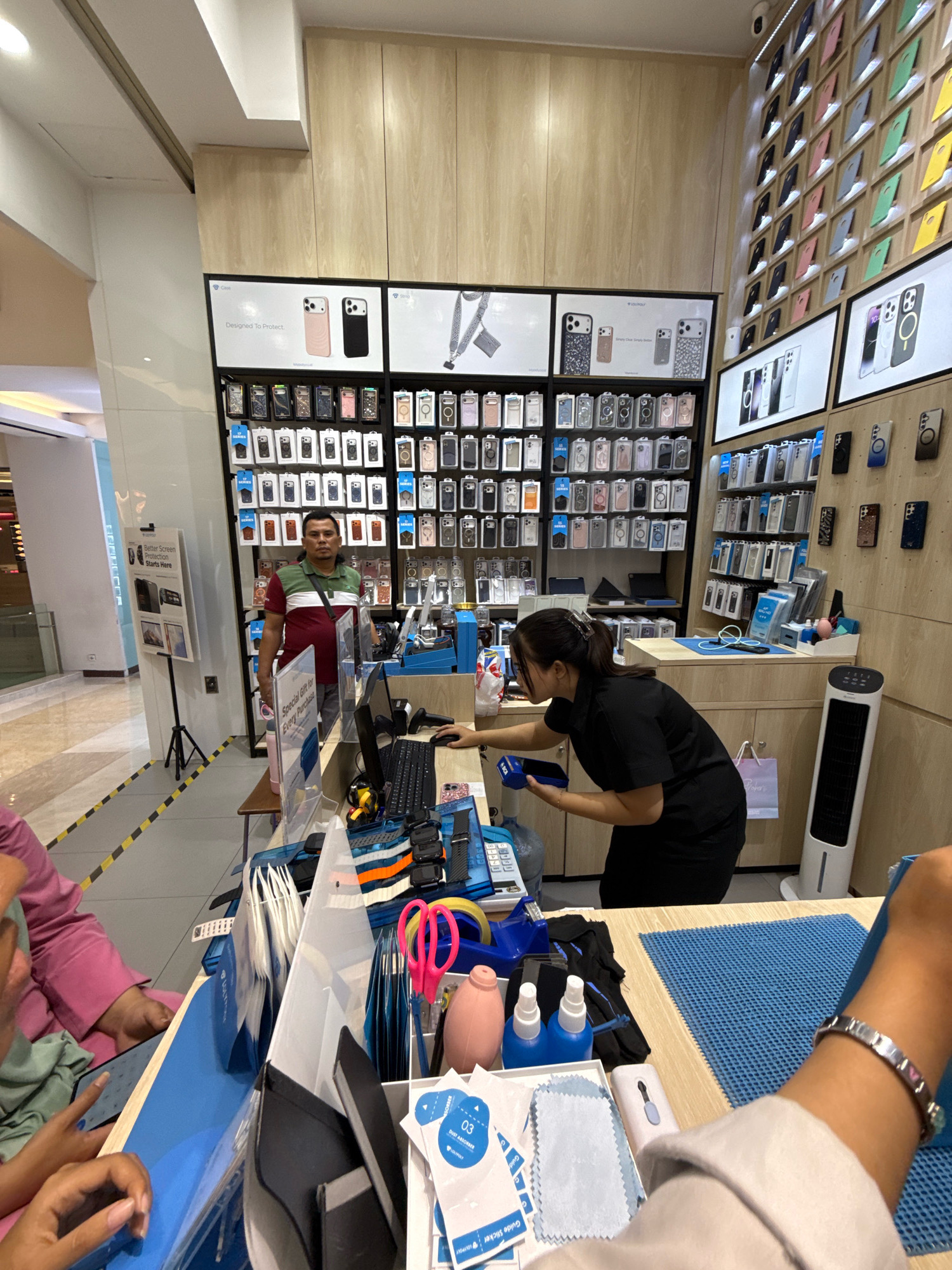 This is a medium-shot photograph taken indoors, likely in a retail store in Surabaya, Indonesia, given the context clues. The scene is a well-lit, organized display of mobile phone accessories.
In the foreground, a counter is laden with various items. A blue mat covers a portion of the counter. To the left, a person's arm, adorned with a silver bracelet, is visible, suggesting they are a customer or employee interacting with items on the counter. Various cleaning cloths, spray bottles, and packets are laid out on the blue mat. A pink-handled pair of scissors and a roll of tape are also present.
Behind the counter, a woman in a black shirt and pants is actively engaged, possibly at a point-of-sale terminal, holding a dark blue object. She appears to be interacting with a computer monitor and a scanner. To her right, a tall, white, vertical air cooler stands against a light wood-paneled wall.
Further back, a man wearing a red, green, and white striped t-shirt and grey pants stands in front of a display. He looks directly at the camera. He has a dark bag slung across his chest.
The background features extensive wall displays filled with numerous colorful mobile phone cases, predominantly for iPhones, organized neatly in rows. Some display panels show product images and branding, such as "Designed To Protect." The overall environment suggests a mobile phone accessory shop. The lighting is bright, illuminating the products and the space effectively.