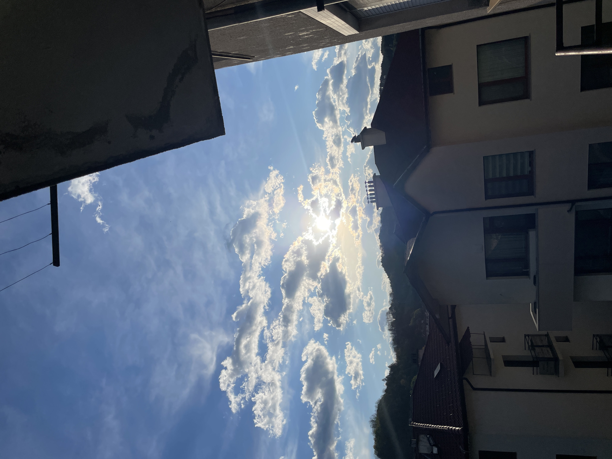 The image shows a view looking up from what appears to be a courtyard or narrow space between buildings in Zlatograd, Bulgaria. A bright, sunny sky is visible, filled with scattered cumulus clouds, and the sun is shining through the clouds, creating a burst of light. The image captures a view that includes the sky, clouds, and parts of adjacent buildings. The buildings are constructed of light-colored material. The scene suggests an urban setting with a combination of natural elements.