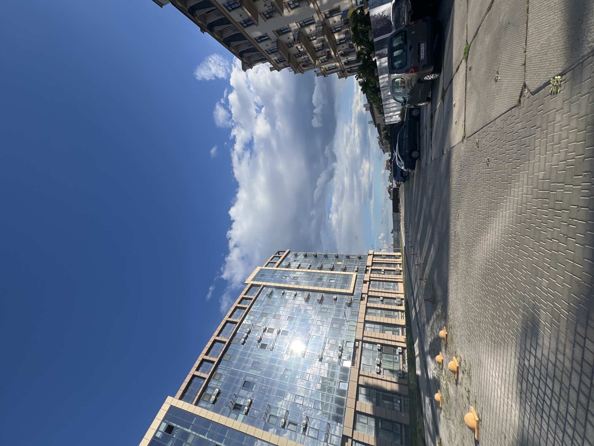 In Fontanka, Ukraine, the image captures a street scene featuring modern and older architectural styles under a partly cloudy blue sky. On the left, a contemporary building with a glass facade reflects the bright sunlight, showcasing multiple air conditioning units. Yellow traffic bollards are visible in the foreground, casting long shadows on the paved surface. To the right, an older, multi-story building with a more traditional design, including balconies and decorative elements, stands. Several cars are parked along the street and on a paved area. The scene is devoid of any people, indicating a quiet moment captured in the urban environment.