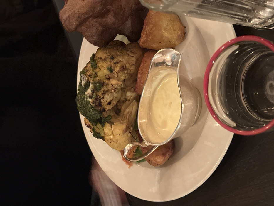 A plate of food featuring a Yorkshire pudding, roasted potatoes, and cauliflower with a green topping. A small silver gravy boat filled with a light-colored sauce sits on the plate. A drinking glass with a red rim is to the right of the plate. The plate is white and the table is dark.