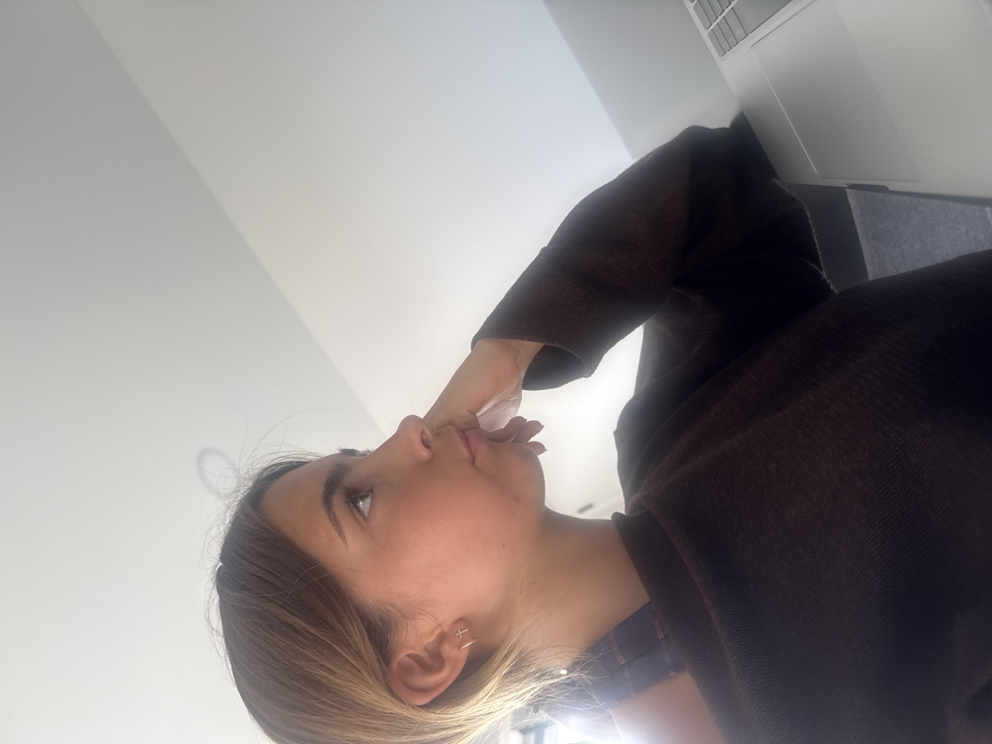 The image shows a woman sitting indoors, angled upwards. She has light brown hair with blonde highlights, fair skin, and is wearing a brown sweater. Her right hand is touching her face near her mouth. She is looking upwards, towards a light source. A laptop is partially visible to the right of the image. The background is a plain white wall. There is no information given to suggest that the image was taken in Torres Vedras, Portugal.