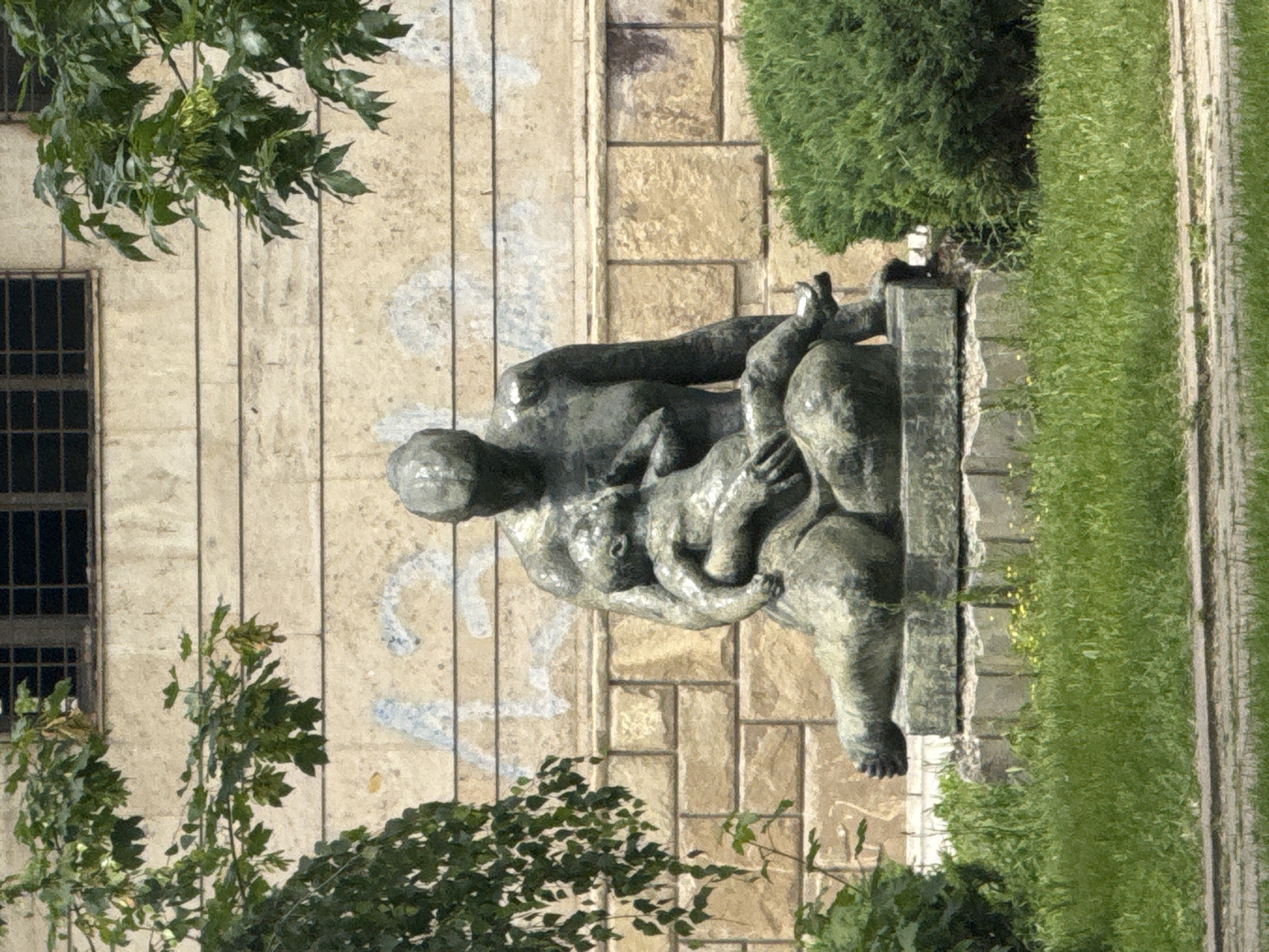 The image shows a statue in Strazhitsa, Bulgaria, situated next to a building with barred windows and graffiti. The statue depicts human figures, possibly a family, in an embrace. Lush greenery surrounds the statue, including trees and grass. A paved walkway borders the grass. The background includes a building facade composed of vertical panels.