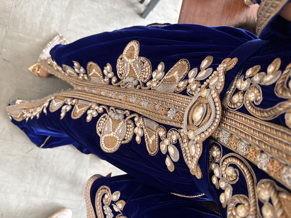 A person is depicted from an elevated perspective, wearing a full-length, royal blue velvet caftan or similar traditional garment. The garment is extensively hand-embroidered with intricate patterns in gold, silver-toned, and light tan threads, featuring stylized floral and abstract motifs. Numerous cream-colored pearls and bead-like embellishments adorn the embroidery, particularly along the central opening and collar. A prominent, ornate central buckle-like element, also embellished with pearls, is visible on the front. Portions of the wearer's feet are visible at the top of the frame; a white shoe is partially seen on the left, and a golden or tan object, possibly footwear, is visible on the right. The person is standing on a light-colored, possibly tiled or stone, floor. A dark brown object is partially visible along the right edge of the frame. The specific location in the city of Paris, France, cannot be determined from the visual information provided within the image.