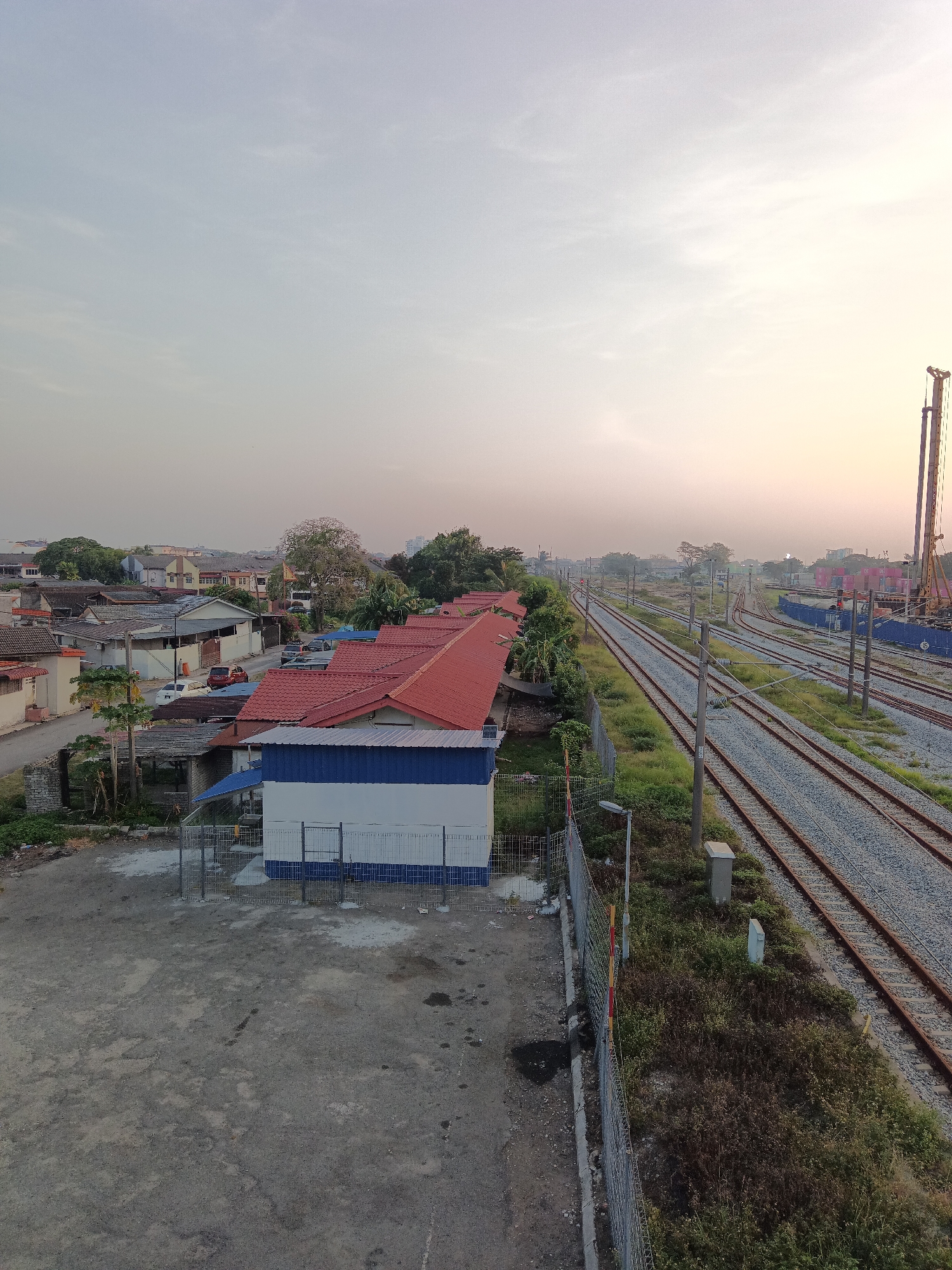 An elevated, wide-angle shot captures a railway yard in Klang City, Malaysia, during the golden hour. The scene is dominated by multiple parallel railway tracks that stretch towards the horizon, flanked by overgrown grass and vegetation. In the foreground, a small, white building with blue trim and a red-tiled roof sits adjacent to the tracks, separated by a metal fence. Beyond this building, a cluster of houses with red-tiled roofs extends into the middle ground, with some parked vehicles visible on a road. Further in the distance, more houses and trees dot the landscape, and a large construction crane is visible on the right side of the frame, suggesting ongoing development. The sky is a soft gradient of pale blue and yellow, indicating either sunrise or sunset. The overall atmosphere is quiet and serene, with no people or animals visible in the frame.