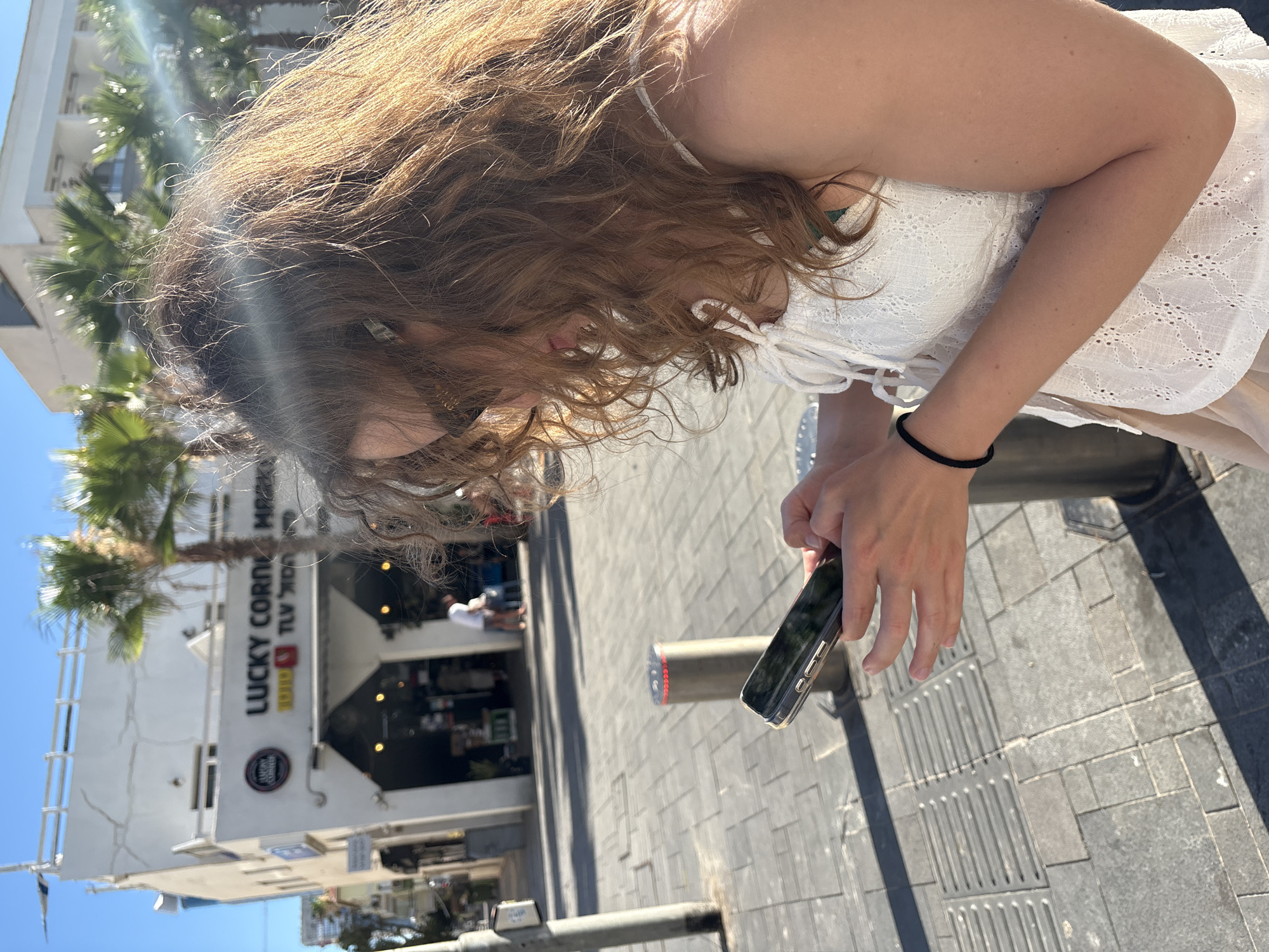 A medium shot shows a young woman with long, curly brown hair and sunglasses looking down at her smartphone, which she holds in her hands. She is wearing a white lace top and a light-colored skirt. Her arm is adorned with a black bracelet. The scene is set outdoors on a sunny day, with a paved sidewalk in the foreground and a building with the signage "LUCKY CORNER MARKET" visible in the background. Palm trees are scattered throughout the scene, suggesting a warm climate. The overall setting is consistent with a street in Tel Aviv, Israel.