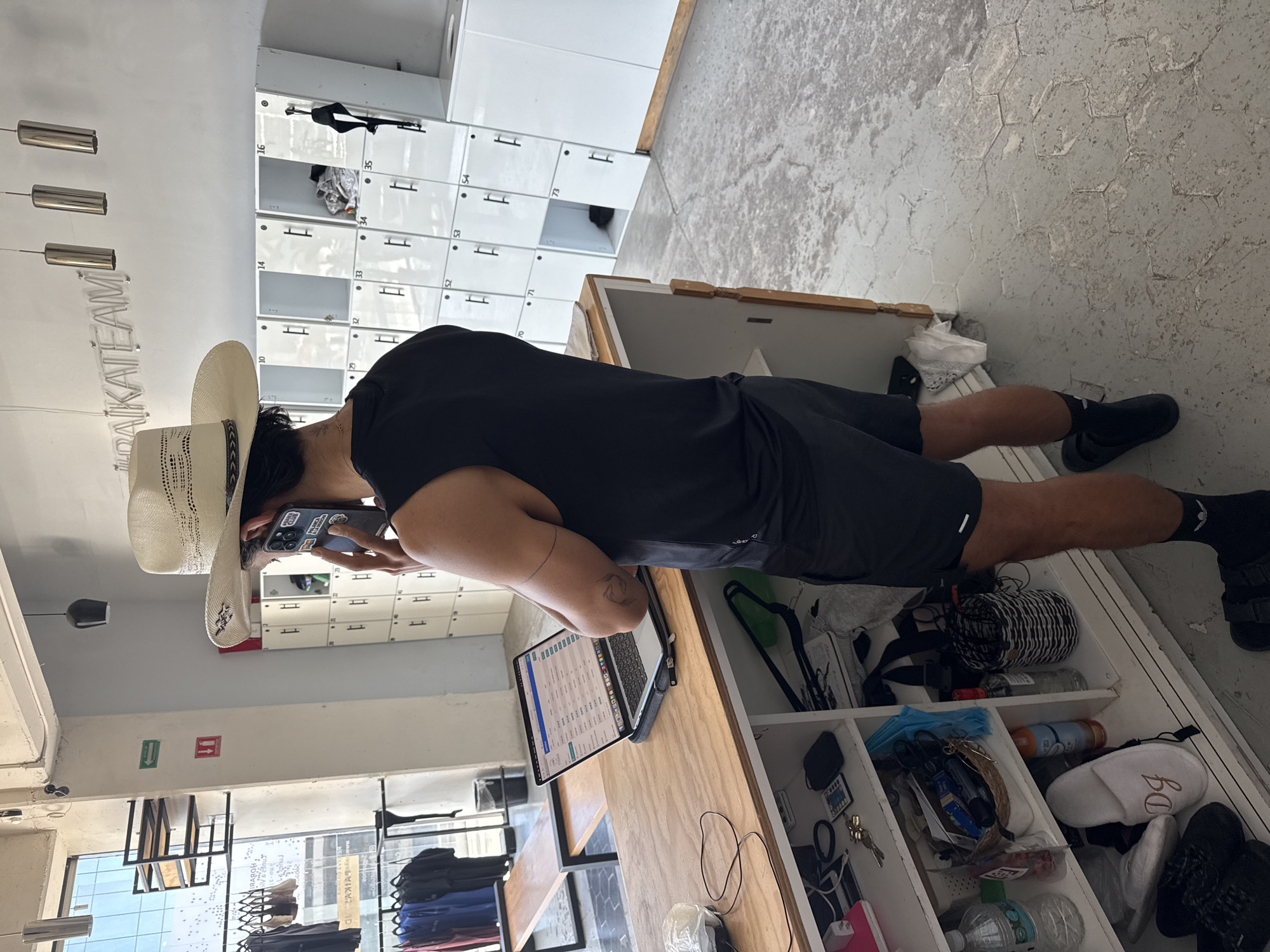 The image captures a person standing inside a bright, modern interior, likely a retail store or a communal space with personal storage. The individual, dressed in a black t-shirt, black shorts, and a straw cowboy hat, is engrossed in their smartphone, holding it up as if taking a photo or video.
A wooden counter runs along the right side of the frame, where an open laptop is visible. Below the counter, a series of open cubbies and drawers reveal various personal items, including a water bottle, coiled black and white rope, and a white slipper with the letters "BD" embroidered on it. In the background, a wall of white lockers, numbered (e.g., 10, 14, 16, 33, 34, 35, 52, 53, 54, 70, 71, 73), signifies a changing area or storage facility. Above, a neon sign displays the word "RAKATEAM."
The floor is a light grey, possibly concrete or tiled with a hexagonal pattern that shows signs of wear. The space is well-lit, suggesting daytime. Through a large window or glass panel on the left, reflections show glimpses of clothing displays and an urban outdoor environment, providing context for the location in Puebla, Mexico. Green and red directional signs are visible on a distant wall. The person's face is obscured, preventing the assessment of their emotions.