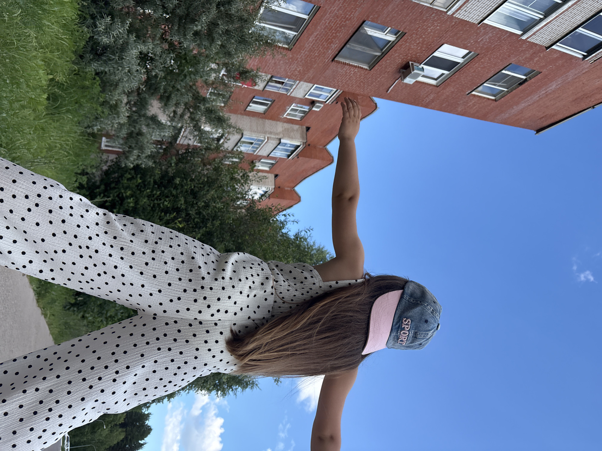 A young girl, seen from behind, stands outdoors wearing a white jumpsuit with black polka dots and a blue baseball cap with "SPORT" embroidered in pink lettering. Her long brown hair falls down her back. She is standing in front of a red brick building with multiple windows and is reaching up with her right arm. The background is a clear blue sky, with some greenery and trees on the left side of the image. The scene appears to be set during the daytime under good weather conditions. The image is oriented vertically with the top of the frame towards the right.