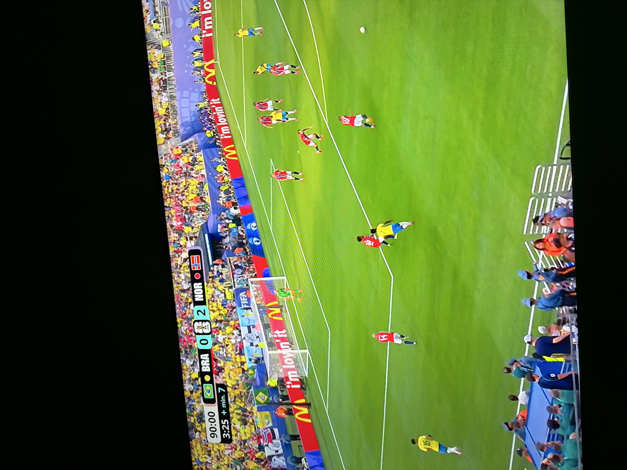 The image shows a soccer match in progress. The score displayed on the screen indicates that Brazil (BRA) has 0 goals and Norway (NOR) has 2 goals. The game time is 90:00, with an additional 7 minutes indicated. Players in red and yellow jerseys are visible on the green field, some actively engaged in the game. Spectators fill the stands in the background, many wearing yellow. Advertising boards with the McDonald's logo and the text "I'm lovin' it" are present around the perimeter of the field. The scene is a stadium setting, implying a public sporting event. There is no information in the image that would suggest the location is Ognianovo, Bulgaria.