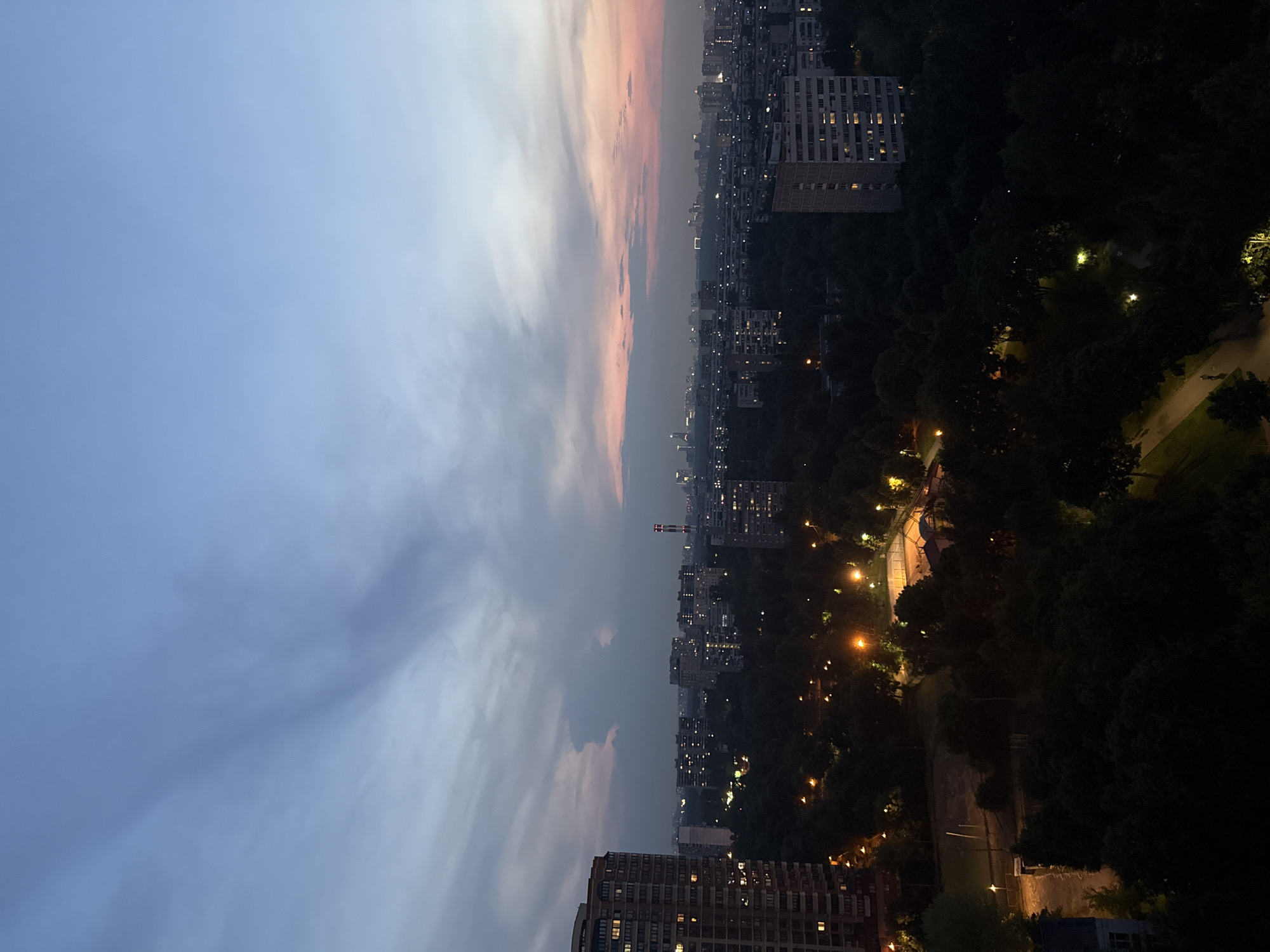 The image depicts a twilight view of a residential area in Moscow, Russia. The sky transitions from a pale blue at the top to hues of pink and orange near the horizon, indicating the end of daylight. Several multi-story apartment buildings are visible, with lights on in many of the windows, suggesting an evening scene. A densely wooded area with mature trees separates some of the buildings. Streetlights illuminate pathways and a sports field within the parkland. The overall atmosphere is one of urban life settling down for the night.