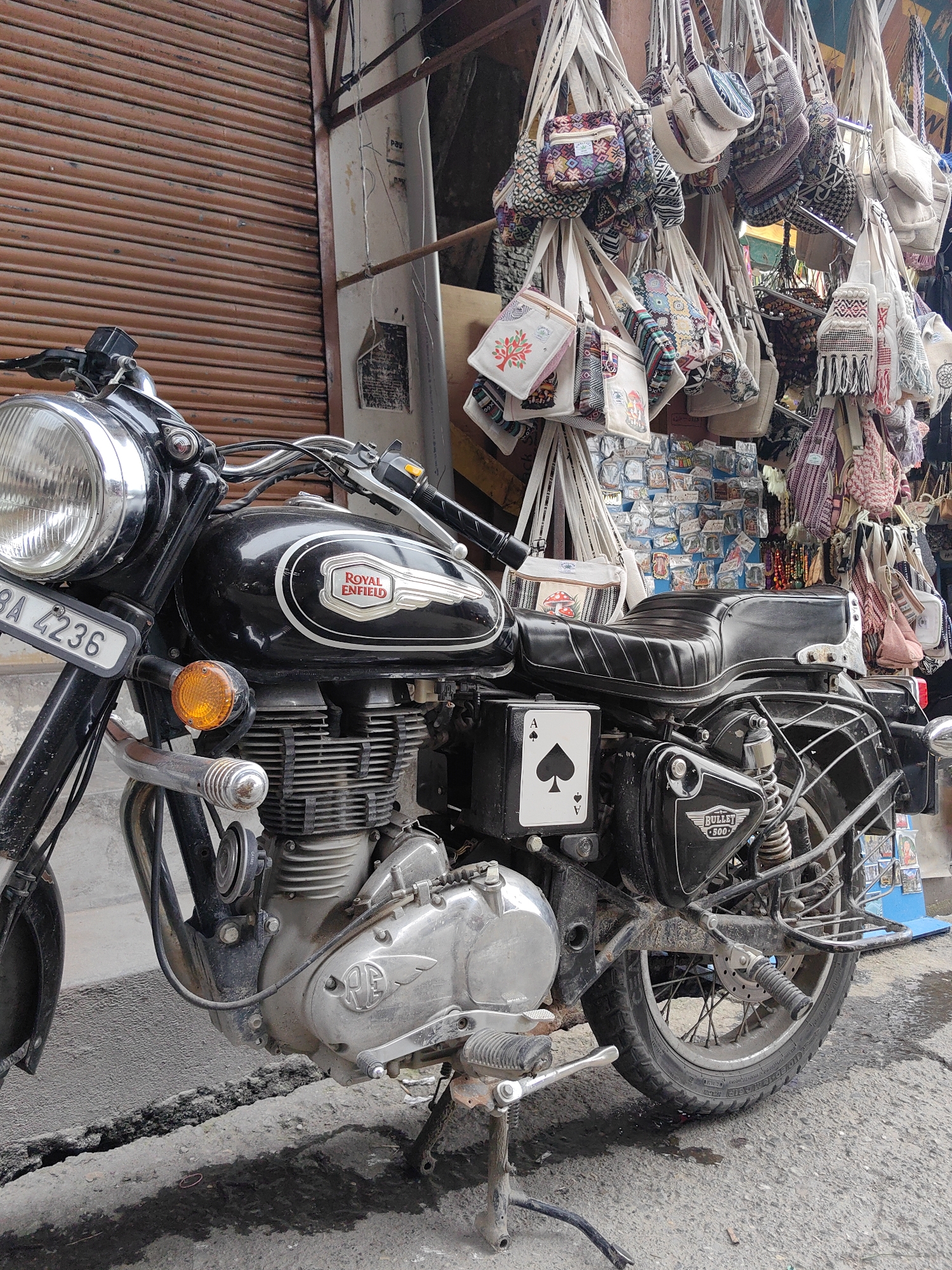 In Vashisht, India, a black Royal Enfield Bullet 500 motorcycle is parked on a dusty, cracked concrete surface with water puddles. The motorcycle features a chrome headlight, a Royal Enfield emblem on its fuel tank, and the Ace of Spades playing card displayed on its side panel. Behind the motorcycle, a market stall is visible, displaying a variety of handmade bags and small items hanging from a metal rack. A brown, corrugated metal shutter forms the background.
