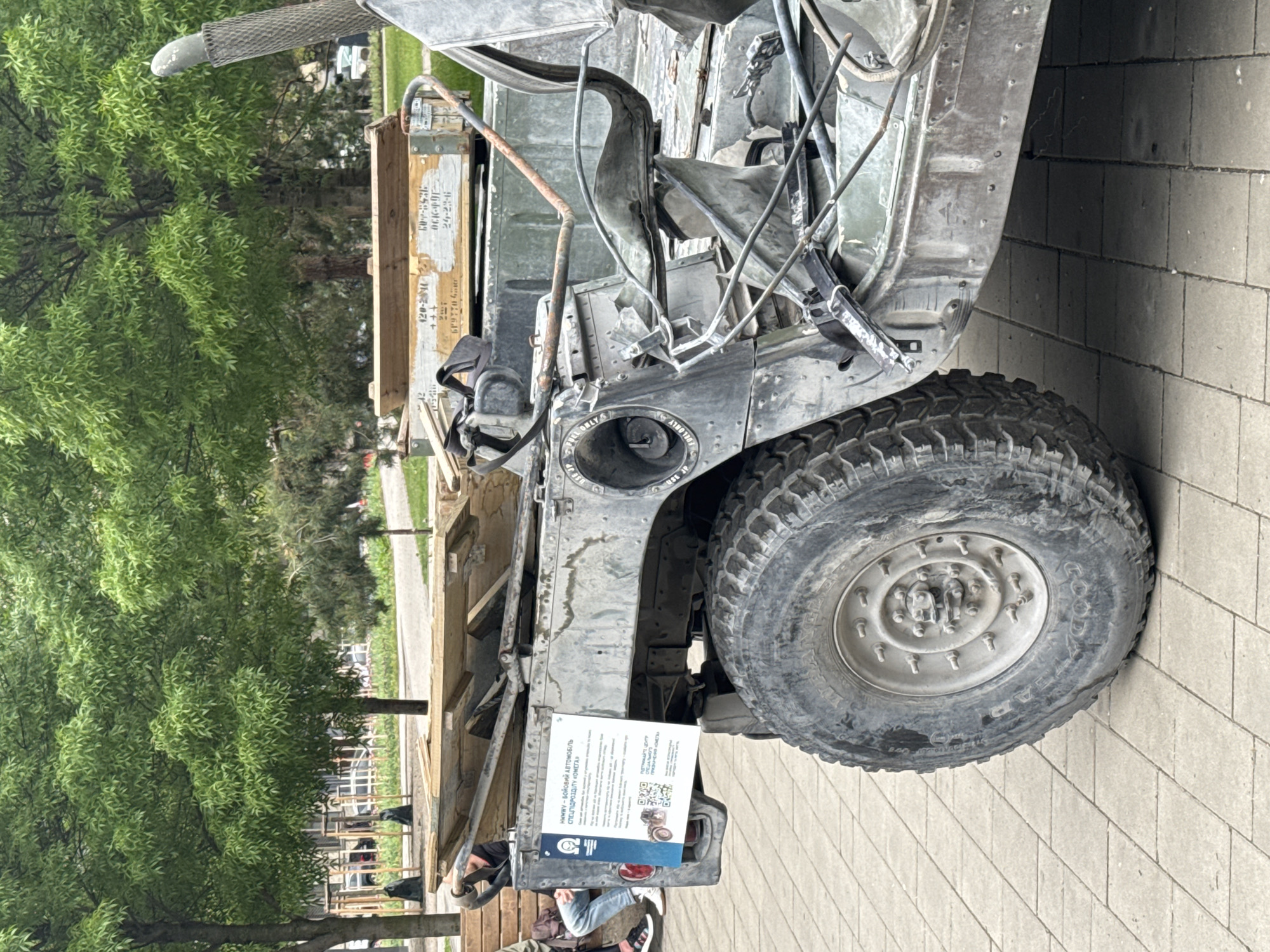 This photograph depicts a partially destroyed military vehicle, possibly a Humvee, on display in an urban setting, likely the city of Kyiv, Ukraine. The vehicle exhibits significant damage, exposing its internal components and wiring. A large tire is visible on the right side of the frame. The vehicle is parked on a paved surface, adjacent to a green area with trees. A sign displaying Ukrainian text and a QR code is attached to the vehicle. Some people are visible in the lower part of the frame, indicating that this is a public display. The overall atmosphere suggests the vehicle is exhibited as a testament to the ongoing conflict.