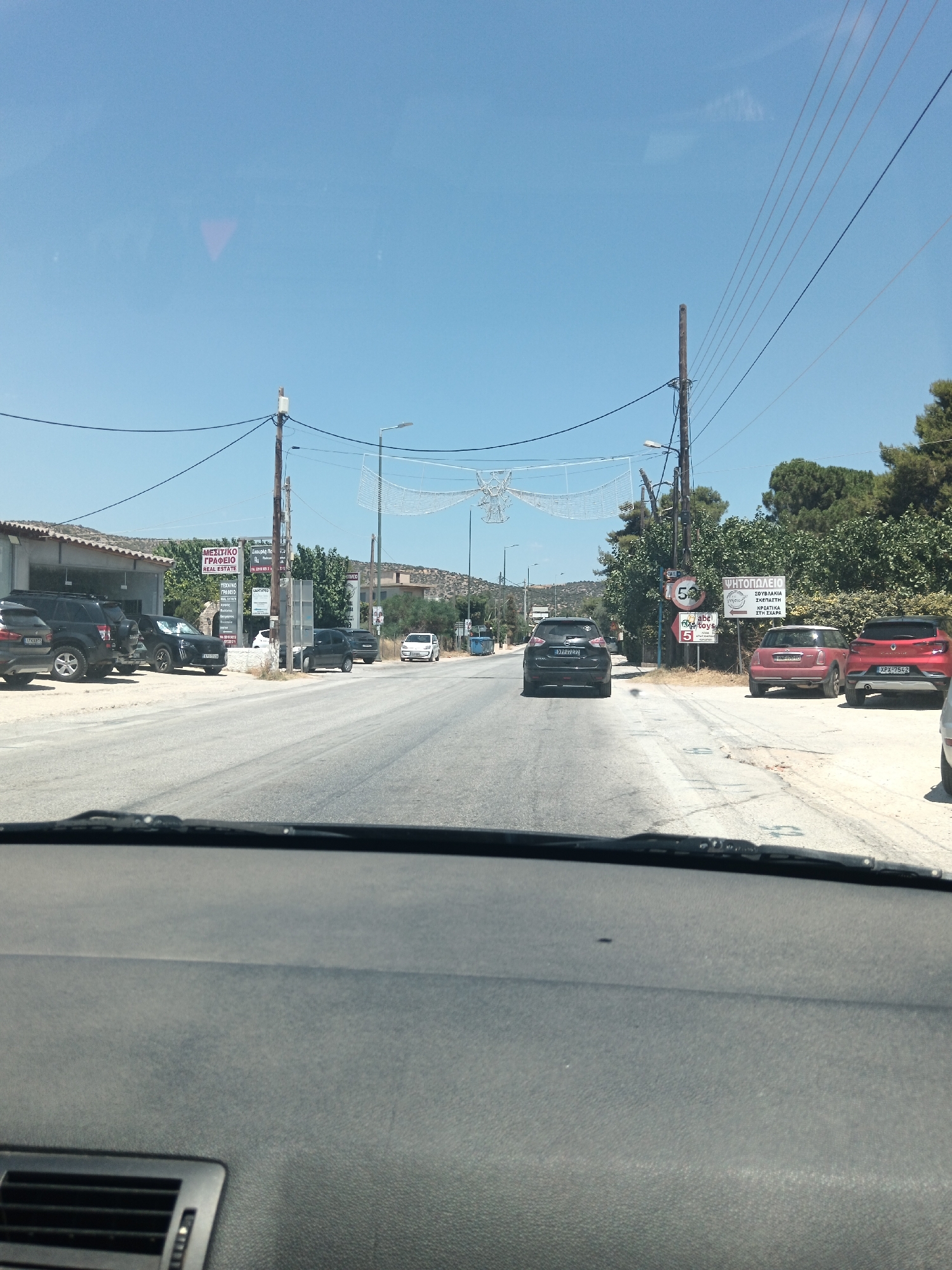 The image captures a street scene in Agia Marina, Greece, on a clear, sunny day. The viewpoint is from inside a vehicle, looking forward. The road is paved and appears to be a two-lane street with parked cars on both sides.
On the left side of the road, several dark-colored SUVs are parked in front of a building with a grey, sloping roof. A sign above the building is visible, with text including "REAL ESTATE" and Greek characters. Further down the street on the left, a black car is parked.
On the right side of the road, a red Mini Cooper and a red Renault Captur are parked. Beyond these cars, there is a dense line of green trees and vegetation. Several utility poles with power lines run along the right side of the street. Signs are visible on these poles, including a circular speed limit sign showing "50", and other signs with Greek text, one of which appears to be for a "PLANT SHOP".
In the middle of the road, a black SUV is driving away from the camera. Further down the street, a white car is visible. The sky is a bright, clear blue. The overall impression is of a quiet, suburban street in a Mediterranean setting.