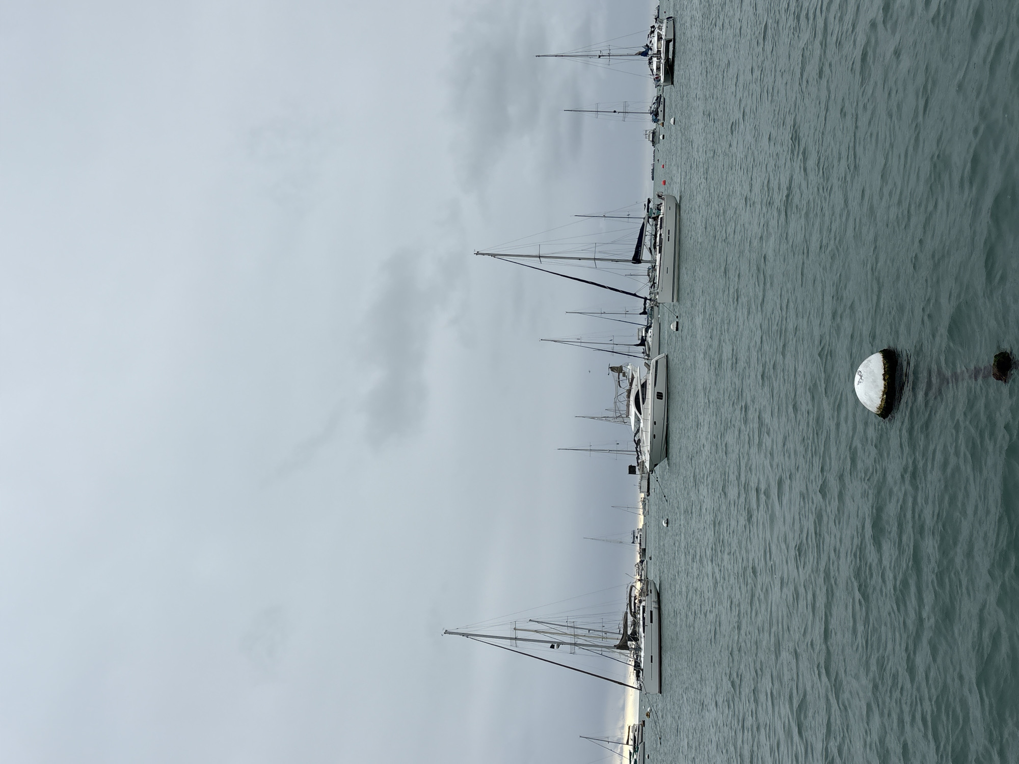 The image depicts a cloudy day with several sailboats and yachts anchored in a harbor. The water is choppy and a buoy is visible in the foreground. In the background, the city of La Punta, Peru, can be seen with buildings and more boats. The sky is overcast with dark clouds, suggesting a possibility of rain. The overall atmosphere is calm and serene, with the boats bobbing gently on the water.