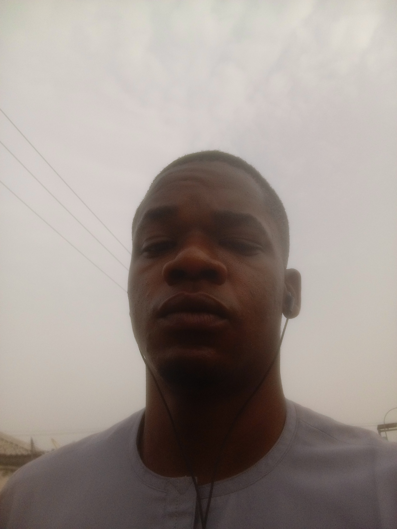 The image is a close-up portrait of a young adult male, likely taken outdoors against a heavily overcast sky. The subject, who appears to be of Black or African descent, is centered in the frame, looking directly at the camera. He has a very short, dark haircut and is wearing a light-colored, possibly white or light grey, crew-neck shirt with a small placket at the neckline. Wired earphones are visible, with one bud in each ear and the wire draped around his neck and chest.
The man's expression is neutral, with his eyes appearing slightly narrowed or partially closed and his lips closed. The lighting is soft and diffused, characteristic of an overcast day, which minimizes shadows and gives the image a somewhat subdued feel.
The background is dominated by a vast expanse of very light grey, almost white, sky. Several thin, dark horizontal lines, resembling power lines or utility wires, are visible in the upper left section of the frame. There are no prominent location cues other than the general outdoor setting and power lines, though the provided context indicates the location is Abuja, Nigeria. No visible text is present in the image.