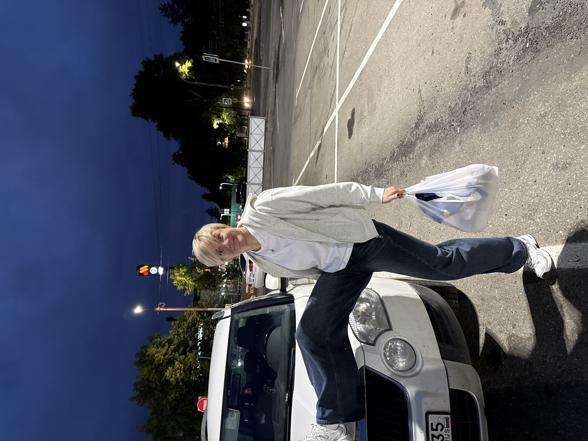 A woman with short blonde hair and glasses stands with her left leg extended onto the hood of a white car. She is wearing a grey hooded sweatshirt, a white t-shirt, and dark blue jeans. In her right hand, she holds a translucent white plastic bag filled with various items. The car is parked in a dimly lit outdoor parking lot. In the background, trees are visible, as well as streetlights and a traffic signal. The sky is dark blue, indicating it is either dusk or nighttime. There are white parking lines painted on the asphalt.