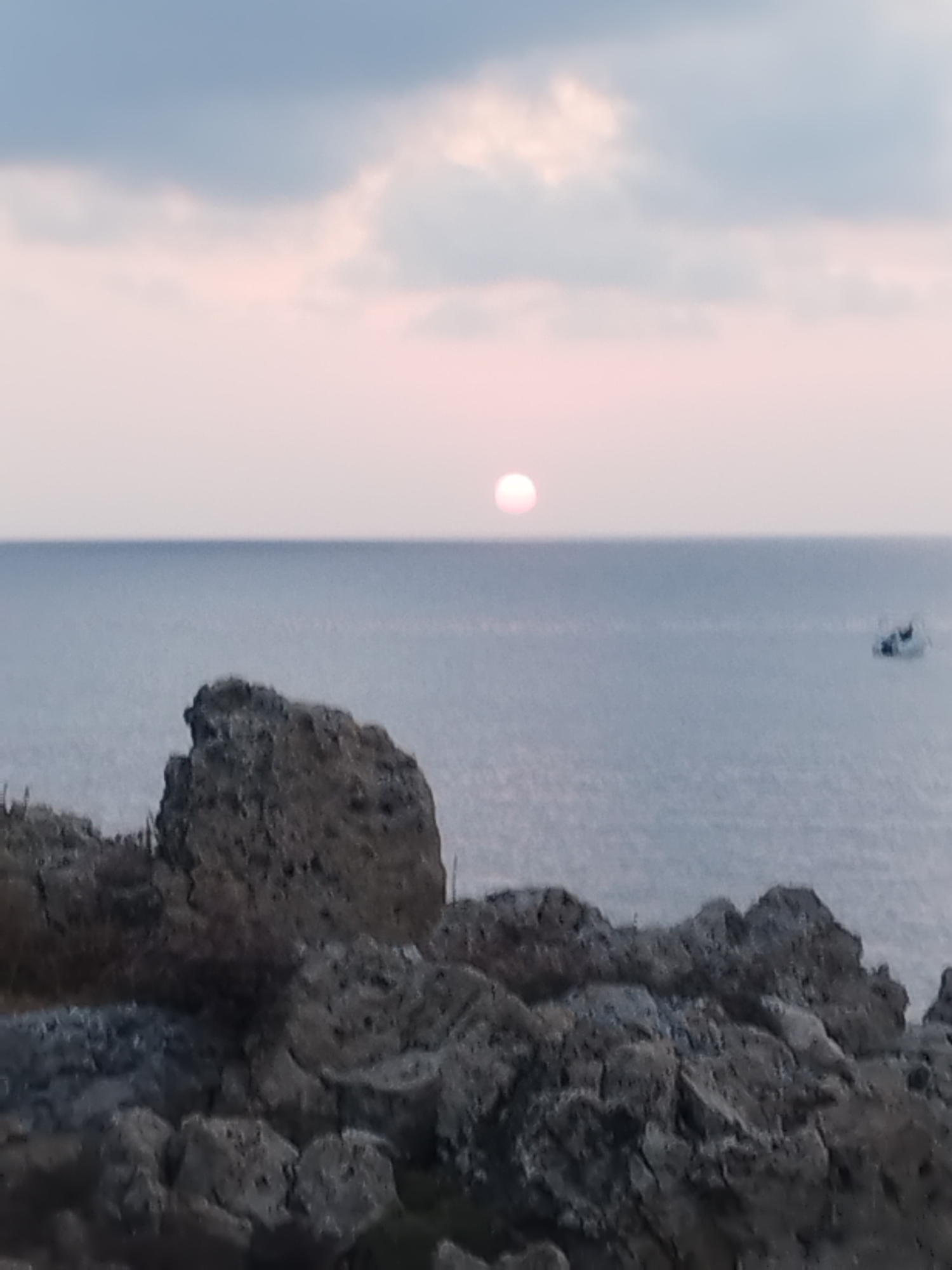 The image shows a serene coastal view at dusk, likely near Antibes, France, given the Mediterranean aesthetic. The foreground is dominated by rugged, dark gray rocks, possibly part of the Cap d'Antibes coastline, with sparse, dry vegetation clinging to them. These rocks create a rough, textured barrier separating the viewer from the sea.
The middle ground features the calm, muted blue waters of the Mediterranean Sea, stretching towards the horizon. A small, white boat with a dark figure on board is visible in the distance on the right side of the frame, suggesting some maritime activity.
The background is defined by the sky, which displays a soft, diffused light characteristic of sunset or sunrise. The sun itself appears as a pale pink, circular orb positioned slightly above the horizon line, casting a gentle glow. The sky transitions from a light pink near the sun to a hazy, pale blue with subtle cloud formations at the top of the image. The overall atmosphere is peaceful and tranquil, capturing a moment of natural beauty.