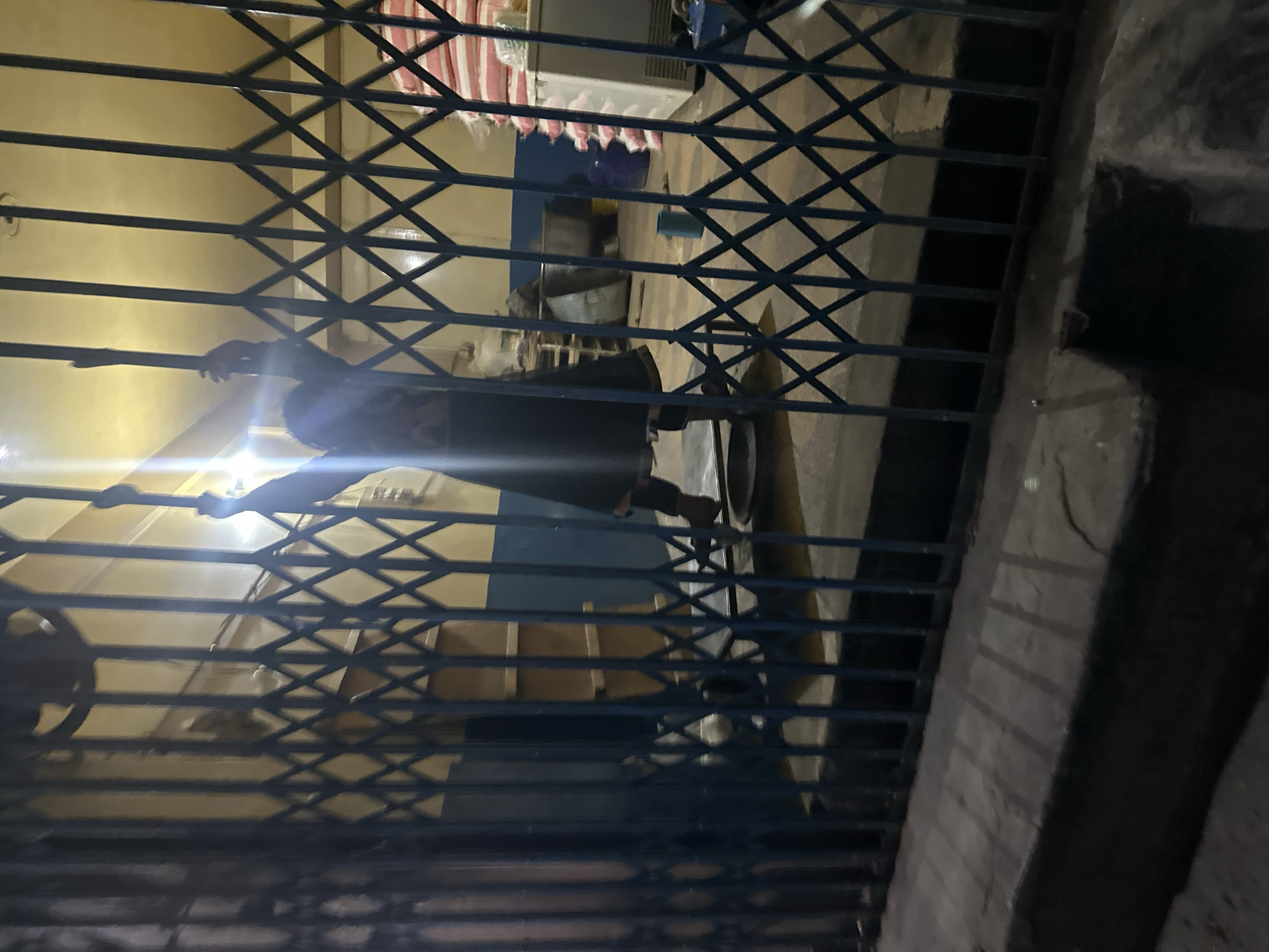The image shows the exterior of a building, as viewed through a blue metal gate. The inside of the building is visible through the gate, and the room has light yellow walls. There are various objects and structures inside, including what appears to be a bed with red and white striped bedding, a refrigerator, and other miscellaneous items. A person stands inside the room, their form partially obscured by the gate. The immediate area outside the gate is concrete. The location is in the city of Unknown, Unknown.