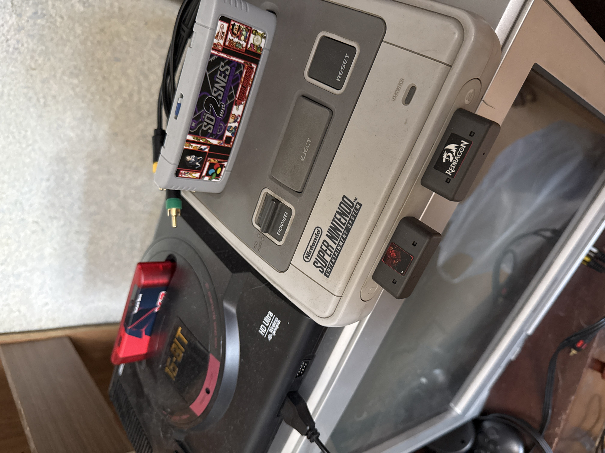 The image shows a stack of retro video game consoles and accessories. On the bottom is a Sega Genesis console with a red game cartridge inserted. On top of the Sega Genesis is a Super Nintendo Entertainment System (SNES) with an SD2SNES game cartridge. Two wireless adapters, labelled "REDRAGON", are attached to the side of the SNES. The consoles appear to be stacked on a table or shelf near a television. The scene appears to be cluttered and indoors. There is no evidence to suggest the location is in Moscow, Russia.