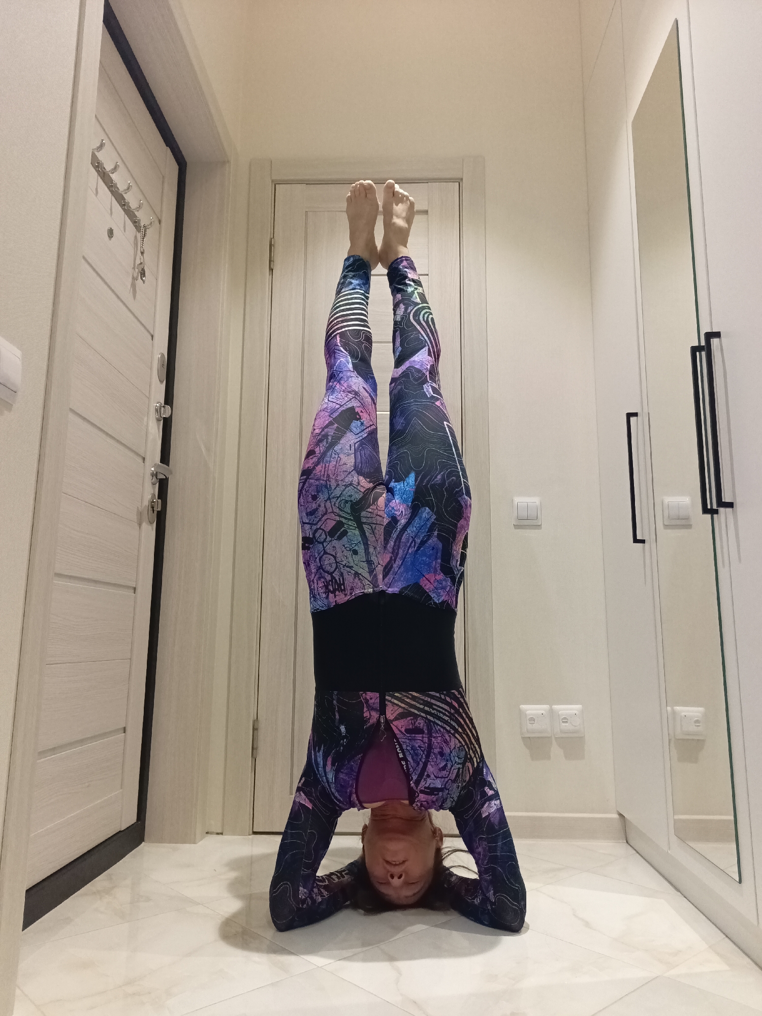A woman performs a headstand in a hallway. She is wearing colorful leggings and a long-sleeved top. Her head is on the floor, and her legs are extended upwards. There is a door to her left and a closet with a mirror to her right. The floor is tiled. The scene is well-lit, with a neutral color palette for the walls and doors.