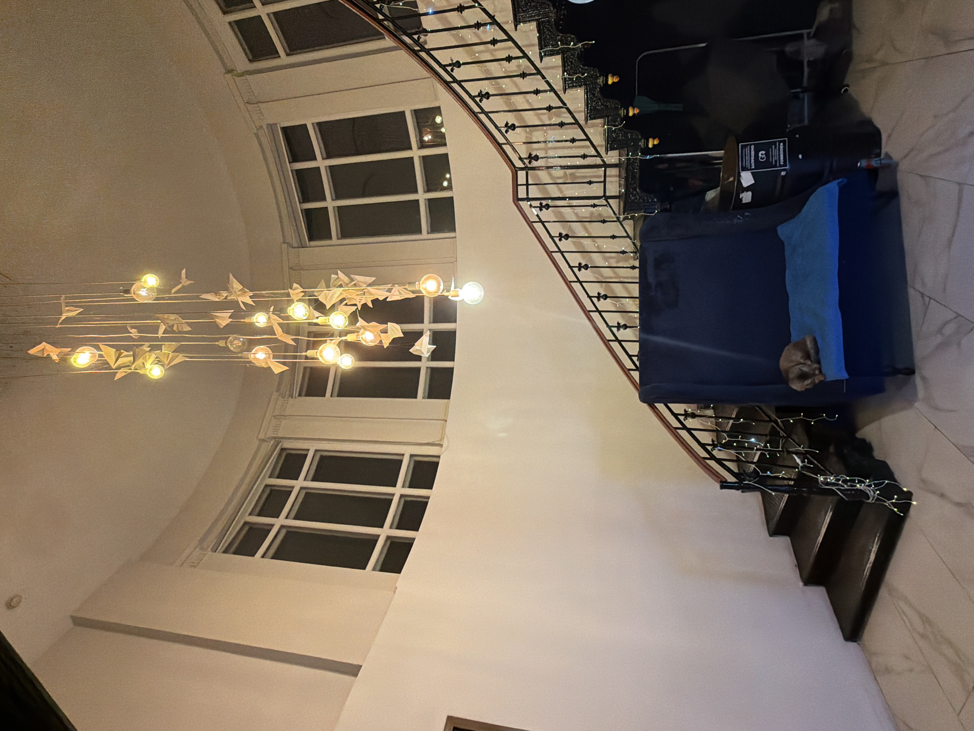 The image depicts an interior scene of a building, likely a hotel lobby or a public space, located in Moscow, Russia. A curved staircase with black wrought-iron railings, adorned with string lights, ascends from the lower level. At the top of the stairs, a large, dark blue armchair is placed, with a light brown cat sleeping on a bright blue cushion on its seat. Above the staircase, a unique chandelier composed of multiple light bulbs and origami cranes hangs from the ceiling. Large, multi-paned windows with white frames are visible on the curved wall behind the staircase. The walls are painted a light cream color, and the floor is tiled with a marble pattern. In the background, several dark figures and objects are visible, including what appears to be a trash can with a sign. The lighting suggests it is evening or indoors with artificial illumination.