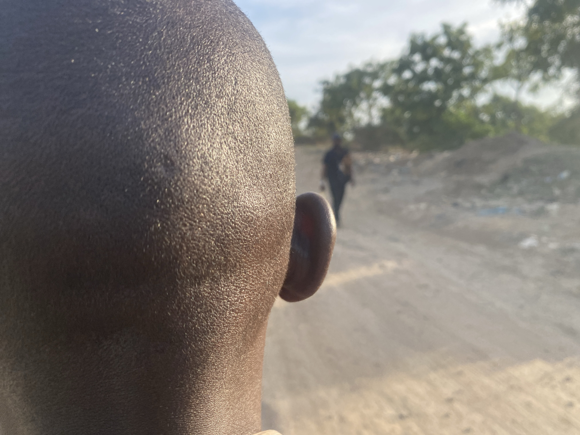 The image presents an outdoor scene in Karmajiji, Nigeria, primarily focusing on the back of a person's head and right ear in the foreground. The individual has dark skin and very closely cropped or shaved hair, illuminated by direct sunlight that creates a noticeable sheen on the skin's surface.
In the blurred background, a dirt path or road recedes into the distance. Another person, a dark silhouette, is visible walking away from the camera along this path. The background also includes indistinct green trees and what appears to be a large pile of dirt or rubble. The sky is bright and clear. The lighting suggests either early morning or late afternoon sun. There is no visible text in the image. The scene conveys a sense of observation or following, set in a semi-rural or developing area.