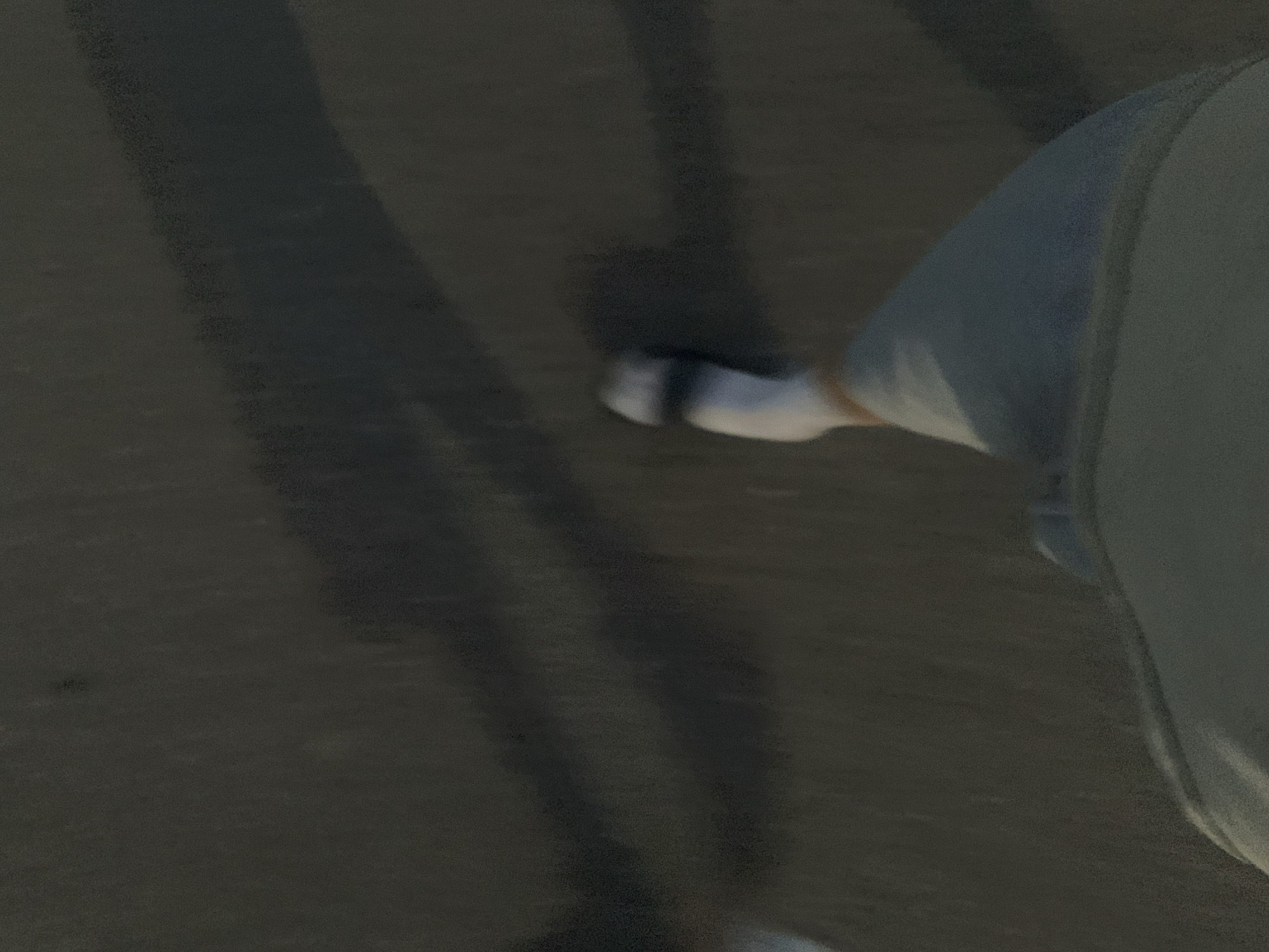 A first-person perspective image shows a person walking on a dark, asphalt surface. The person is wearing a light-colored sock and a denim garment that appears to be a skirt or shorts. Shadows are cast on the asphalt, suggesting an overhead light source. The lighting conditions are dim. The scene is not identifiable as being in Minsk, Belarus.