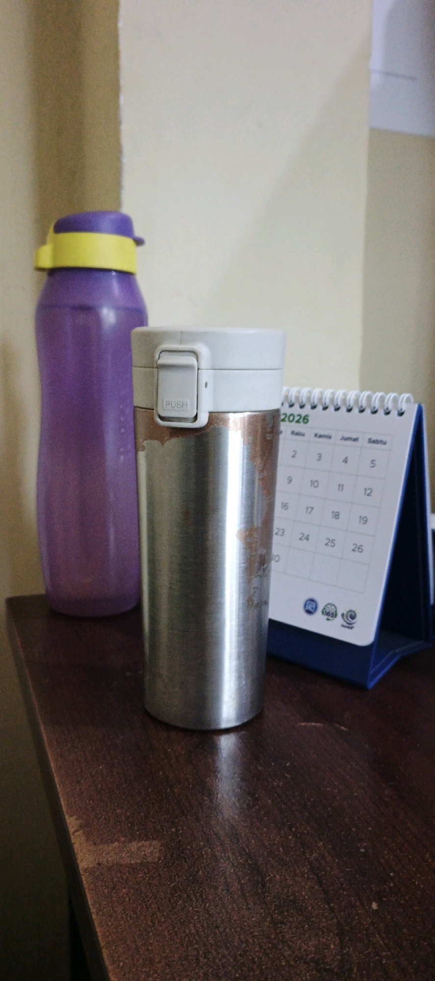 In Tg Kupang, Indonesia, a close-up shot captures a portion of a wooden desk with two objects and a desk calendar. To the left, a tall, purple water bottle with a yellow lid stands upright. To its right, a metallic silver thermos with a white lid is positioned. The thermos has a small grey button with "PUSH" printed on it. A desk calendar, partially visible to the right of the thermos, displays the year "2026" and a grid of dates. The background consists of a light-colored wall. The scene is lit, suggesting an indoor setting.