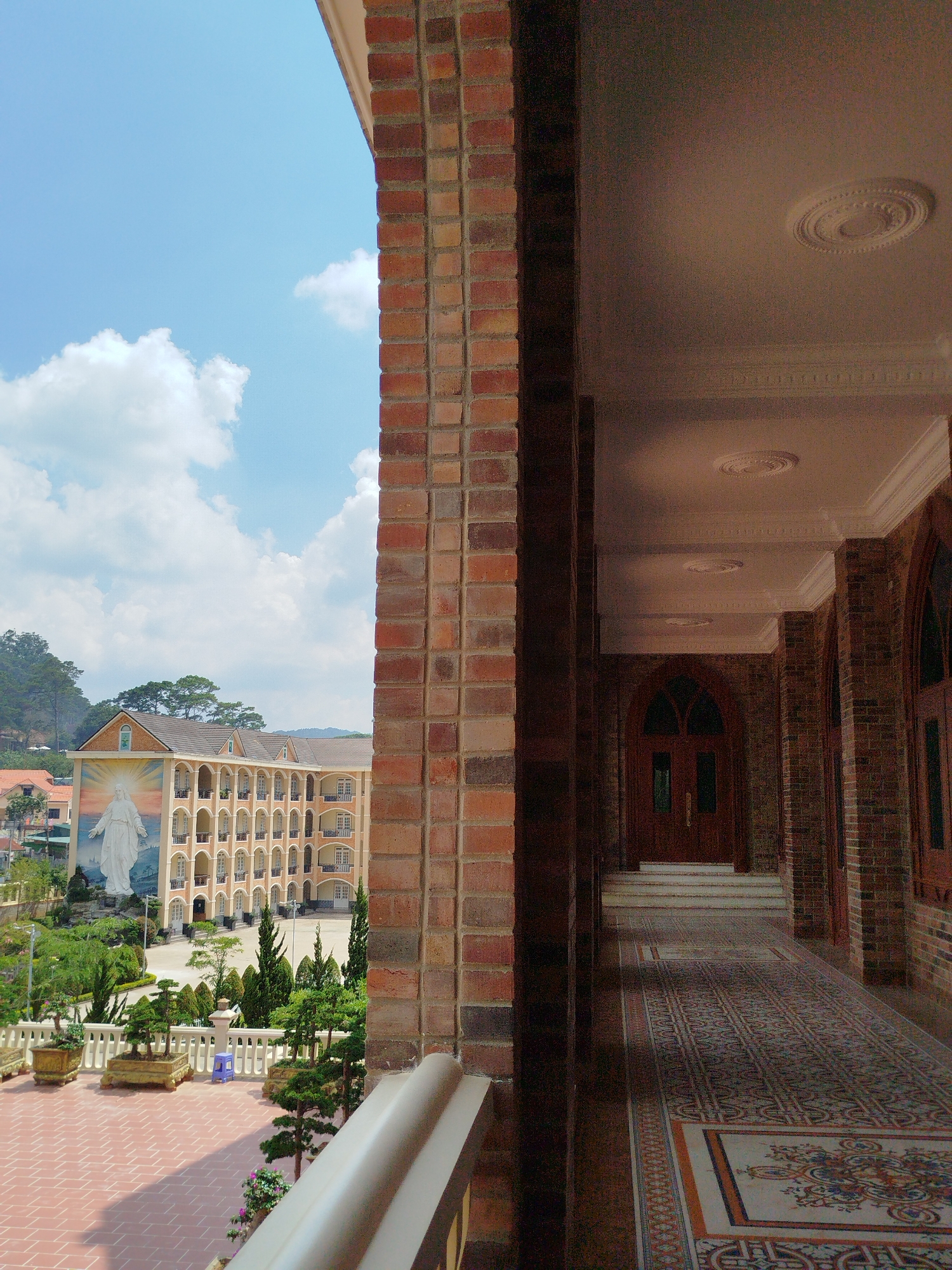 The image depicts a view from a balcony in Cam Ly - Đà Lạt, Vietnam. The balcony has a low, white railing and overlooks a courtyard with potted plants. In the distance is a large building with an image of a religious figure painted on its side. The sky is blue with white clouds. To the right of the frame is a covered walkway with decorative ceilings and tiled floors. A dark wooden door is visible at the end of the walkway. The architecture consists of brick and tile elements, with decorative ceiling rosettes. The scene is overall serene and bright.