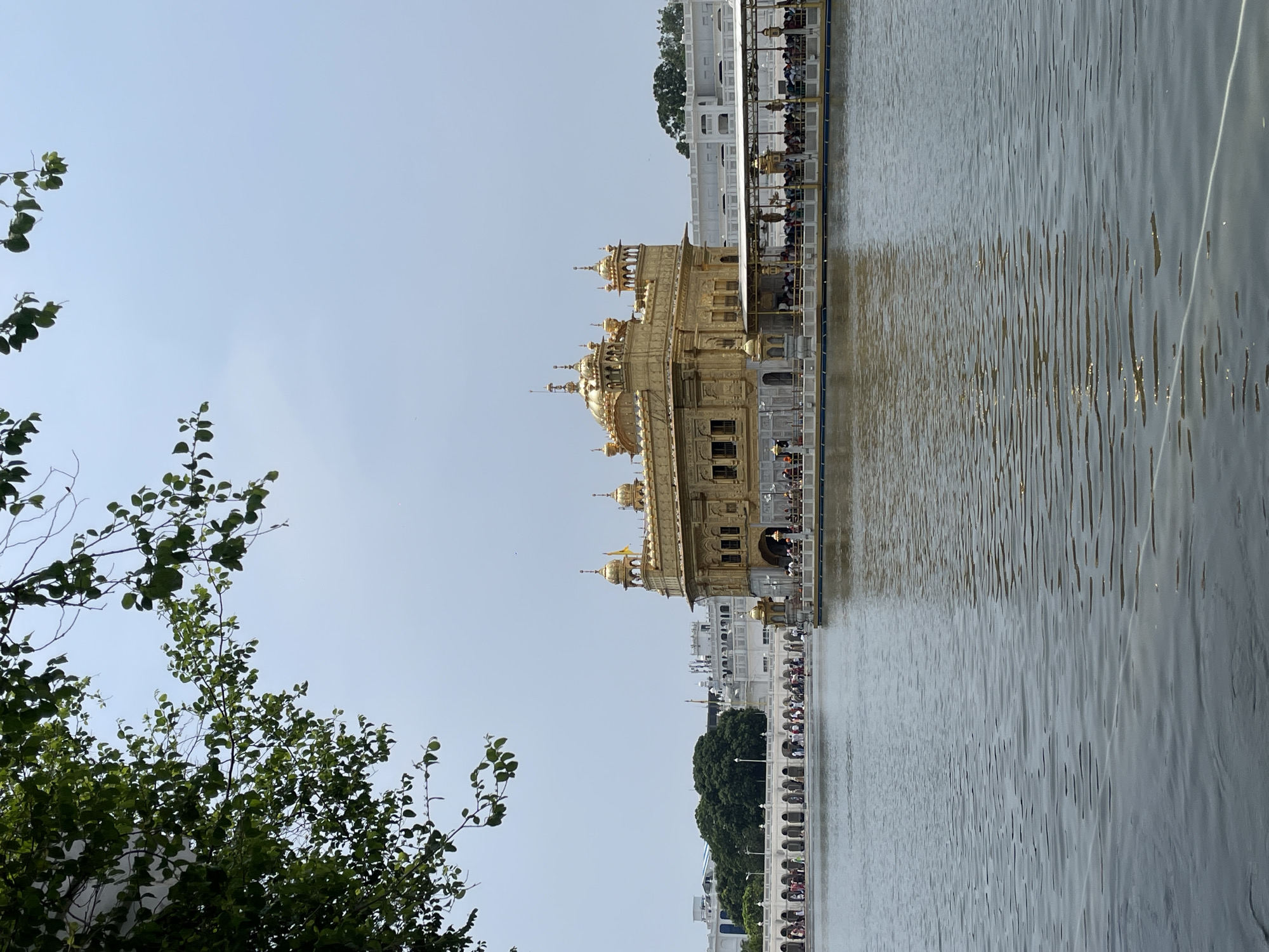 The image depicts the Golden Temple in Amritsar, India, viewed from across a body of water. In the foreground, the water is calm with ripples and reflections of the sky and the temple. The Golden Temple, a prominent Sikh Gurdwara, stands in the center of the image, its golden facade gleaming under a bright sky. The temple complex is surrounded by a walkway and an archway structure with many people visible walking along it. The background shows additional white buildings and trees, with the sky being overcast and a light grey. Lush green foliage from a tree in the upper left corner frames the scene. The overall impression is one of a serene and spiritual atmosphere, with a significant number of visitors present.