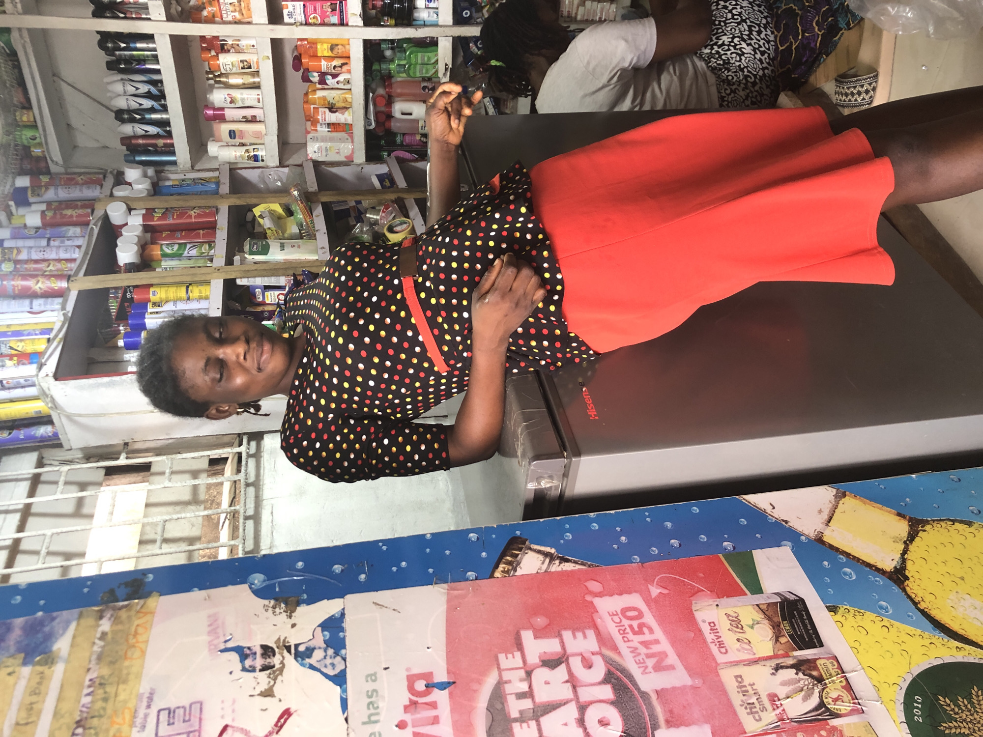 In Ado, Nigeria, a young woman stands leaning against a silver Hisense refrigerator in what appears to be a small convenience store or kiosk. She is wearing a black, short-sleeved top with a red and yellow polka dot pattern, and a vibrant red, knee-length skirt. Her eyes are closed, and her head is tilted downwards, suggesting a moment of rest or contemplation. Her right arm is bent, with her hand resting on the refrigerator, while her left arm is also bent, with her hand positioned near her stomach.
Behind her, shelves are packed with various products, including what seem to be toiletries, drinks, and food items. The store environment is somewhat cluttered, with goods visible on multiple shelves extending upwards. To the left of the woman, a large, colorful poster is partially visible, featuring text and graphics, including what looks like an advertisement for "Chivita Ice Tea." The poster appears to be adhered to a blue, textured surface. In the background, another person, possibly a woman, is seated and facing away from the camera, also appearing to be in the store. The lighting suggests an indoor setting, possibly with ambient light from the store's interior.