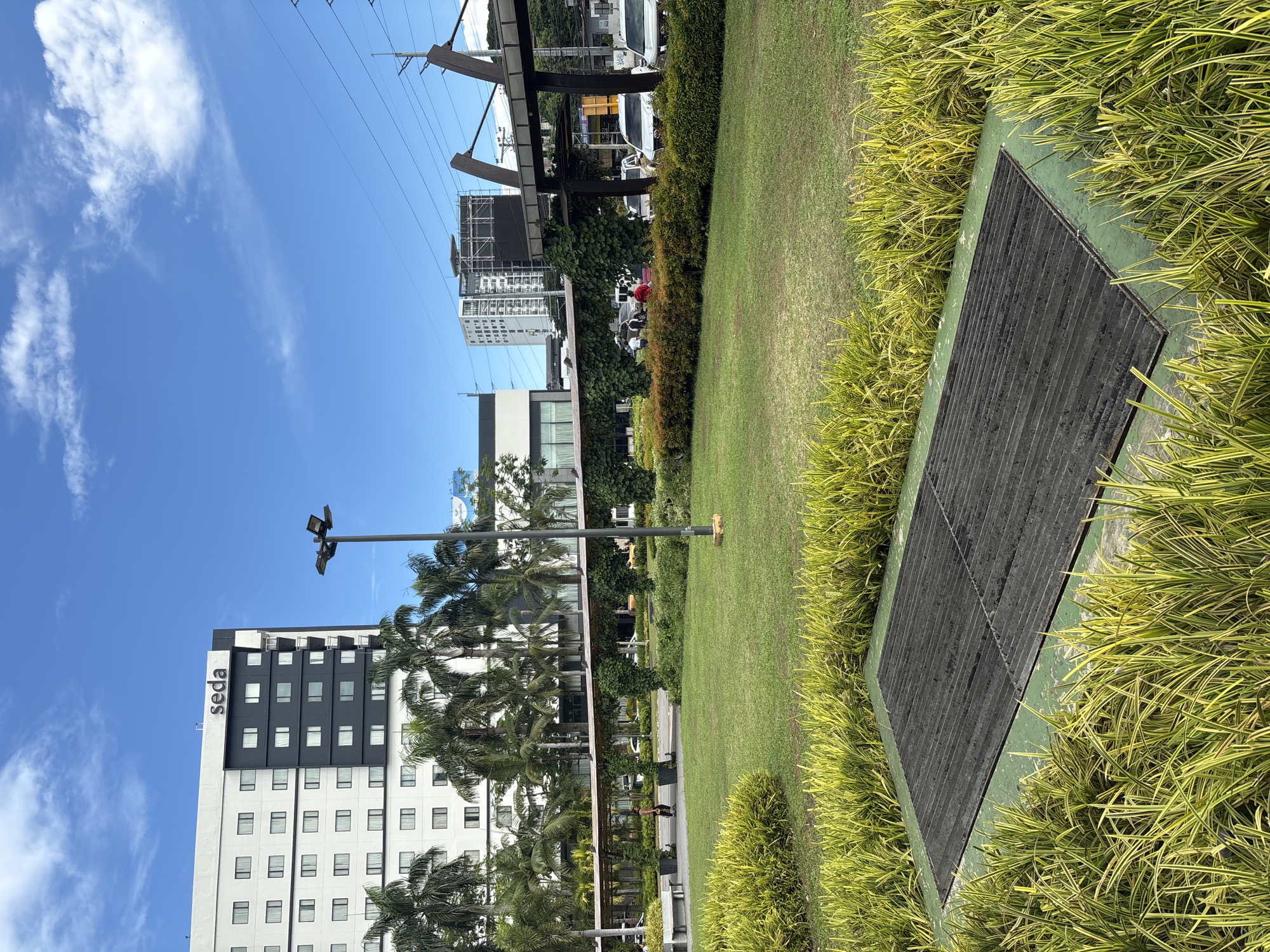 The image captures an outdoor scene in Davao City, Philippines, likely in a commercial or business district. In the foreground, a patch of vibrant green grass is bordered by a bed of variegated ornamental grass. Centered in this grass is a rectangular metal grate, possibly an access point for underground utilities or a drainage cover.
Behind this, a well-maintained lawn stretches towards a covered walkway adorned with climbing vines and punctuated by intermittent trees. Several individuals are visible walking along or near this walkway, one wearing a red shirt. Palm trees are scattered throughout the midground, their fronds reaching towards the sky.
Dominating the background is a modern, multi-story hotel building with a white facade and dark accents, bearing the "seda" logo at its top. Beyond the hotel, more buildings and infrastructure, including what appears to be an elevated walkway or bridge structure, are visible, along with traffic on a road and utility poles with power lines. The sky above is a clear blue, dotted with scattered white clouds.