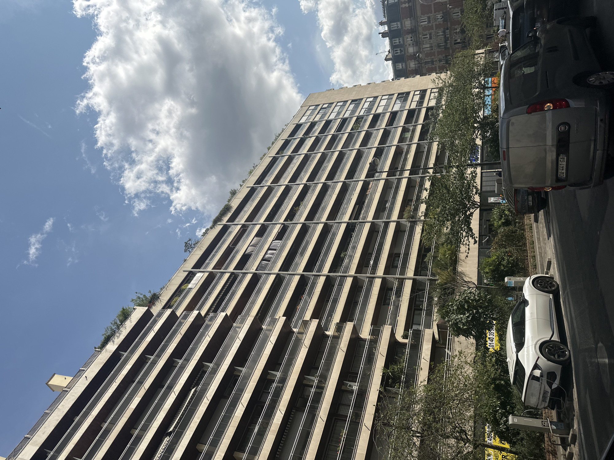In a city scene in Paris, France, a modern, light-colored electric car is parked and charging at a charging station on a street lined with trees. The car is a Kia EV6, distinguished by its distinctive taillight design. Behind the car, a tall, multi-story apartment building with numerous balconies stretches across the frame. The building has a beige facade and appears to be a residential structure. The sky above is a mix of blue with scattered white clouds, suggesting fair weather. Further in the background, other buildings and vehicles can be seen, indicating an urban environment. The overall impression is a street view within a bustling European city.