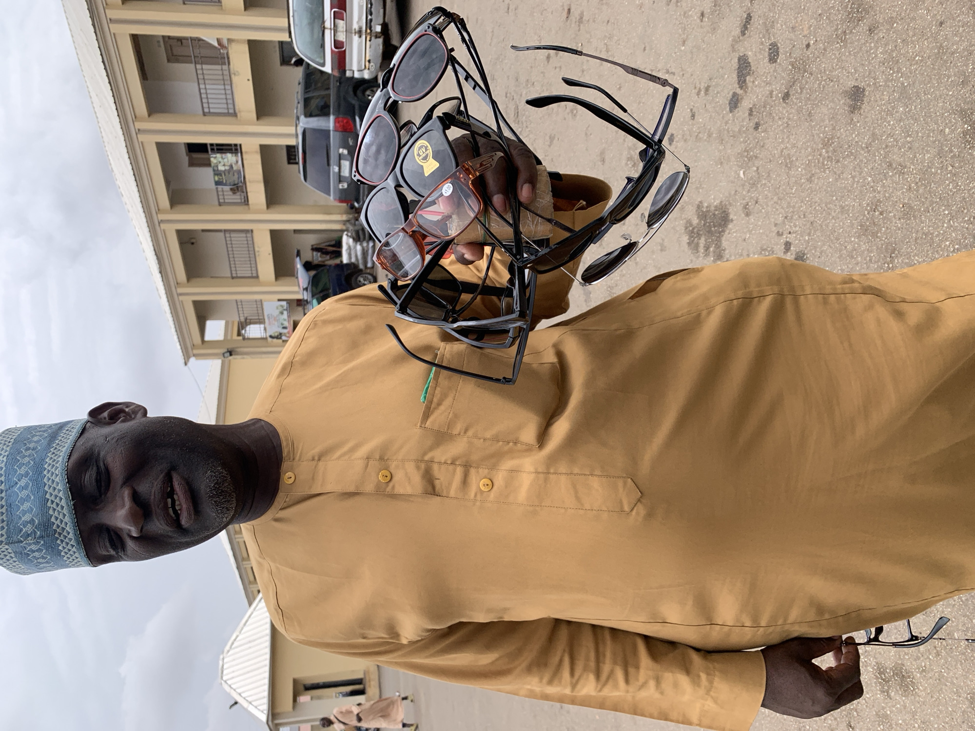 A man stands on a paved surface in Bukuru, Nigeria, holding a bundle of assorted sunglasses in his left hand and a pair of eyeglasses in his right. He wears a light brown tunic and a blue patterned kufi cap. The background features a two-story building with multiple levels of balconies and parked vehicles. The sky is overcast.