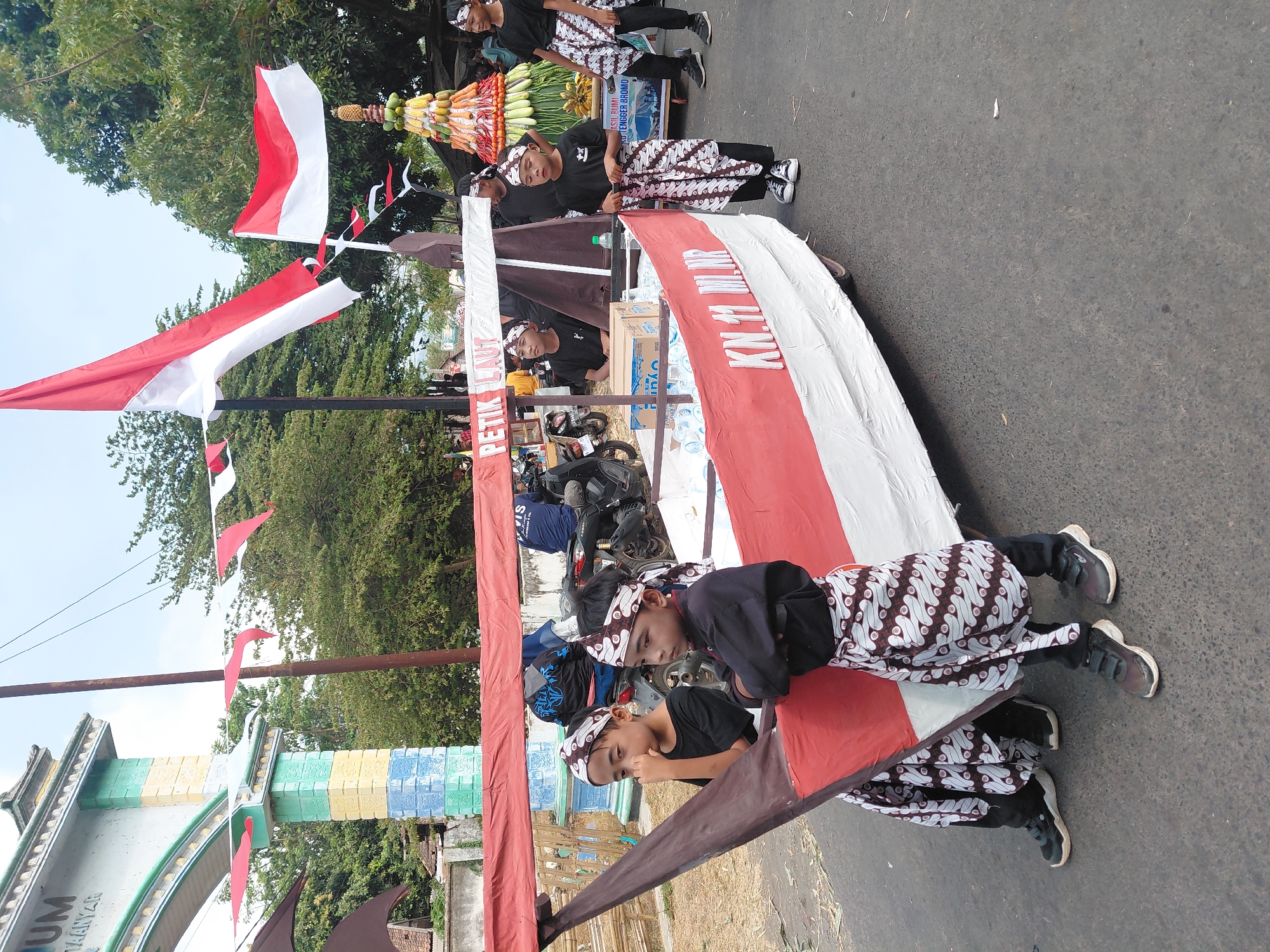 The image depicts a street scene in Bima, Indonesia, likely during a parade or cultural event. In the foreground, a group of young boys, dressed in traditional Indonesian attire including headbands and sarongs with batik patterns, are participating. They are positioned around a large, boat-shaped float decorated in red and white, symbolizing the Indonesian flag. The float has text that reads "PETIK LAUT" and "KN.11 ML.NP". Behind the float, a vibrant, tiered pyramid of fruits and vegetables is visible, adorned with an Indonesian flag on top. The event appears to be taking place outdoors, with trees and buildings in the background. The overall atmosphere suggests a festive and community-oriented gathering.