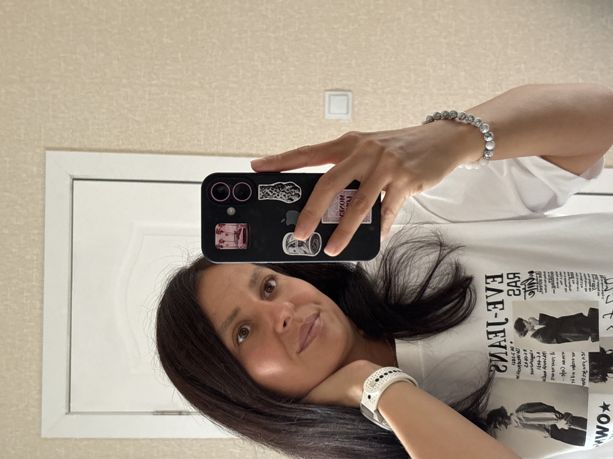 In a close-up, indoor mirror selfie, a woman with dark, shoulder-length hair and brown eyes is positioned in the center of the frame. She is wearing a white t-shirt with black text and graphics, a white digital watch on her left wrist, and a bracelet with white and grey marbled beads on her right wrist. The woman is holding a black smartphone in her right hand, which is covered in various stickers. She is looking towards the phone's camera, with her left hand touching her neck. Behind her, a white door and a textured, beige wall are visible. The scene appears to be set in a residential interior. The location is identified as Chaikovsky, Russia.
