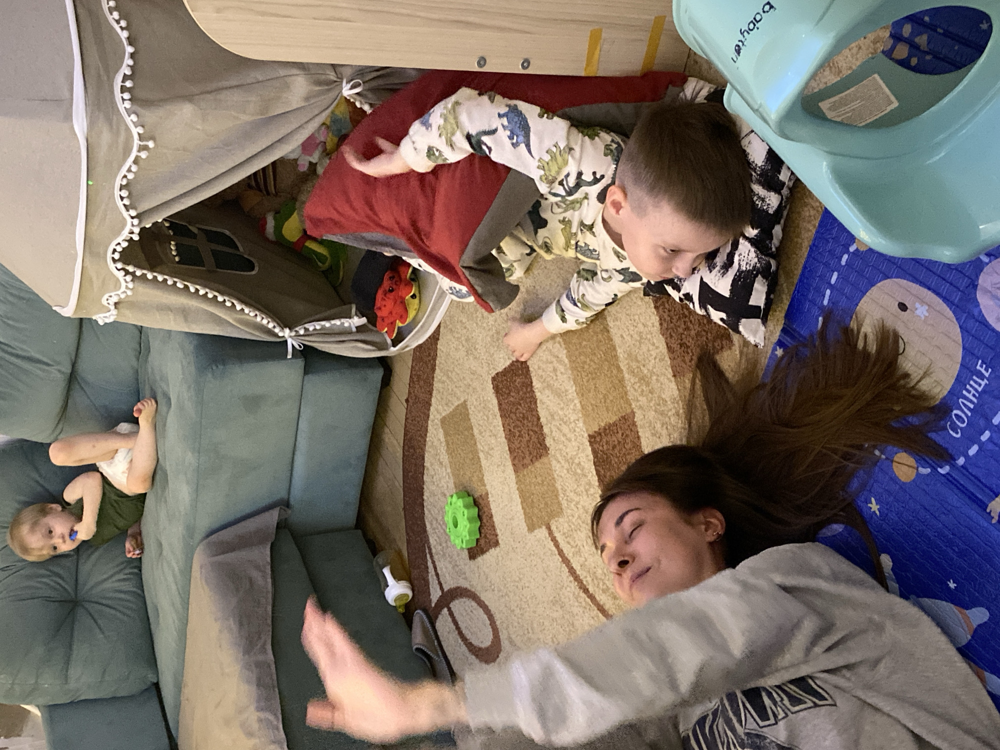 A residential interior scene, likely in Votkinsk, Russia, presents three individuals: an adult female and two young children. The female, with long brown hair, lies on a carpeted floor, her eyes closed. She is wearing a light-colored sweatshirt. A toddler, dressed in dinosaur-patterned pajamas, is next to her on the carpet. A smaller child, wearing a diaper and green top, is lying on a blue couch. A play tent with white trim sits partially under a wooden table. Various toys are scattered around, including a green gear-shaped toy and a baby potty seat. A blue play mat with illustrations is also visible.