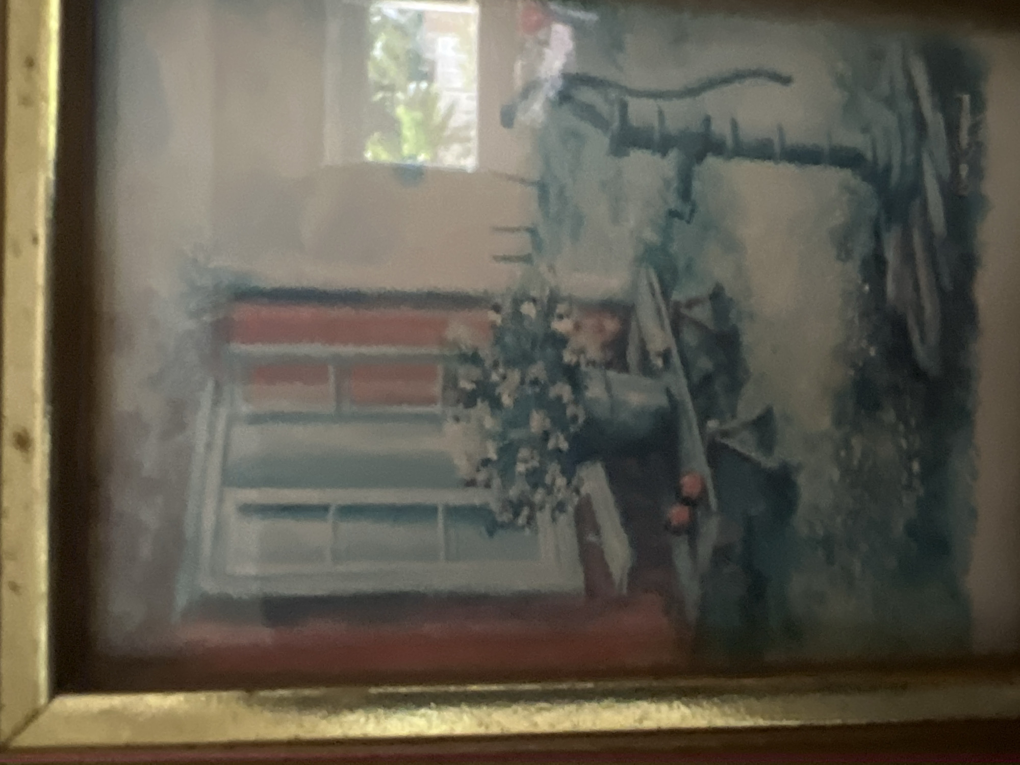 The image displays a framed painting. The frame is gold-toned with a visible inner reddish border. The painting depicts an outdoor environment, situated in the city of Casablanca, Morocco. On the left side of the canvas, a structure features a white-framed window with closed red shutters, set into a reddish-brown wall. A light-colored, possibly wooden, table or bench is positioned beneath the window. Upon this surface rests a grey, metallic-appearing container filled with numerous small white flowers. Adjacent to the container are two distinct, spherical, red objects. The right side of the painting is dominated by a dark, vertical hand pump, exhibiting a curved handle or spout on its right side. The ground area around the pump and table is rendered with a mix of blue and green hues, suggesting foliage or a garden. In the upper right background, a rectangular opening, potentially a window or doorway, reveals a blurred view of green vegetation beyond. No human figures are present, and no actions or interactions are depicted.