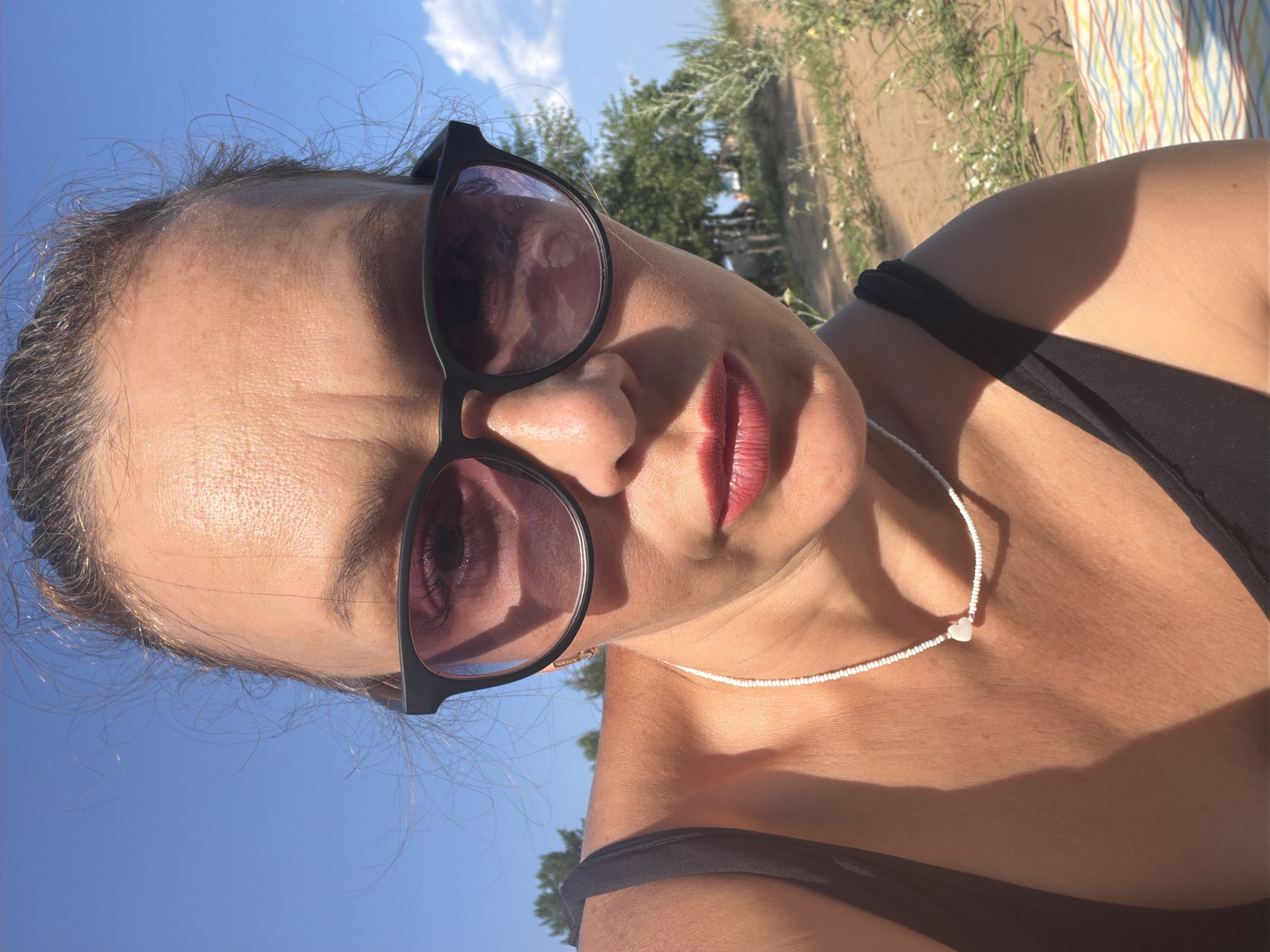 A woman is taking a selfie outdoors on a sunny day. She is wearing sunglasses and has red lipstick on. She is also wearing a black swimsuit and a white beaded necklace with a heart-shaped pendant. The background shows sand, grass, trees, and a blue sky with some clouds. The image does not contain enough information to determine the location in Noviy, Russia.
