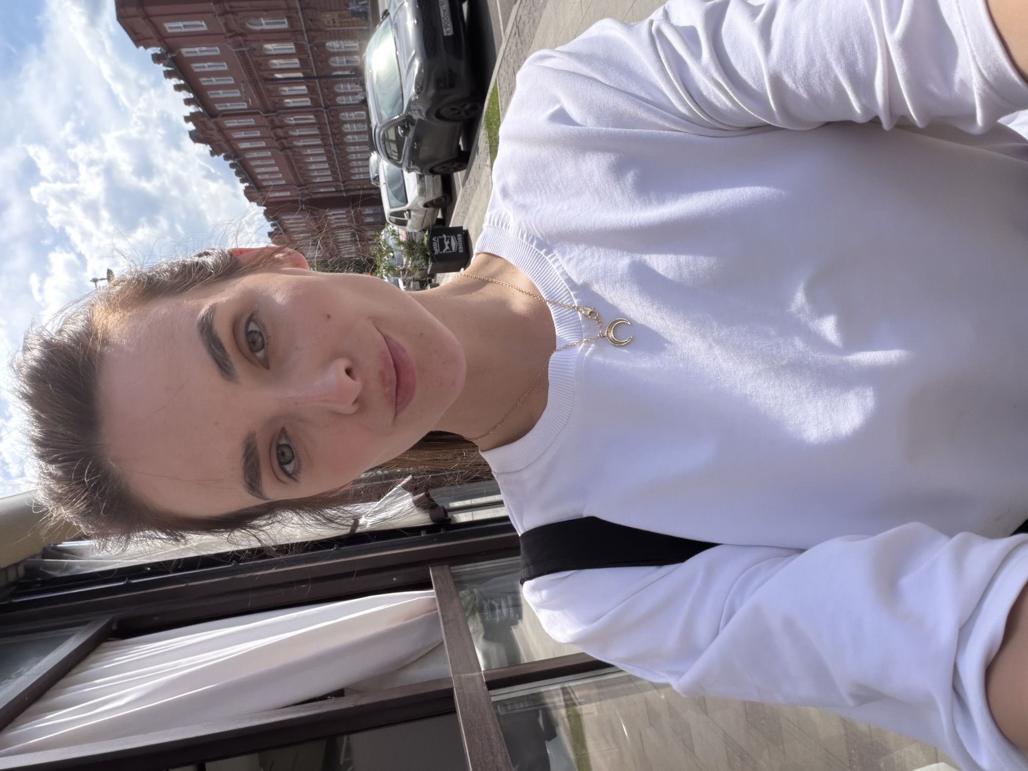 The media file is a selfie of a young woman, taken outdoors during the daytime. She is wearing a white t-shirt and a thin gold necklace with a crescent moon pendant. The background shows a classic red brick building with arched windows and an overcast sky with some clouds. A dark car and a lighter colored vehicle are parked on a sidewalk in front of the building. There is also a black trash bin visible. The woman's face is partially visible, and she is smiling slightly. She has light brown hair and light-colored eyes. The scene appears to be a street in Ufa, Russia, based on the location context. The lighting suggests it is daytime, likely late morning or afternoon, with the sun intermittently breaking through the clouds. The overall mood is casual and relaxed. No visible text is present in the image.