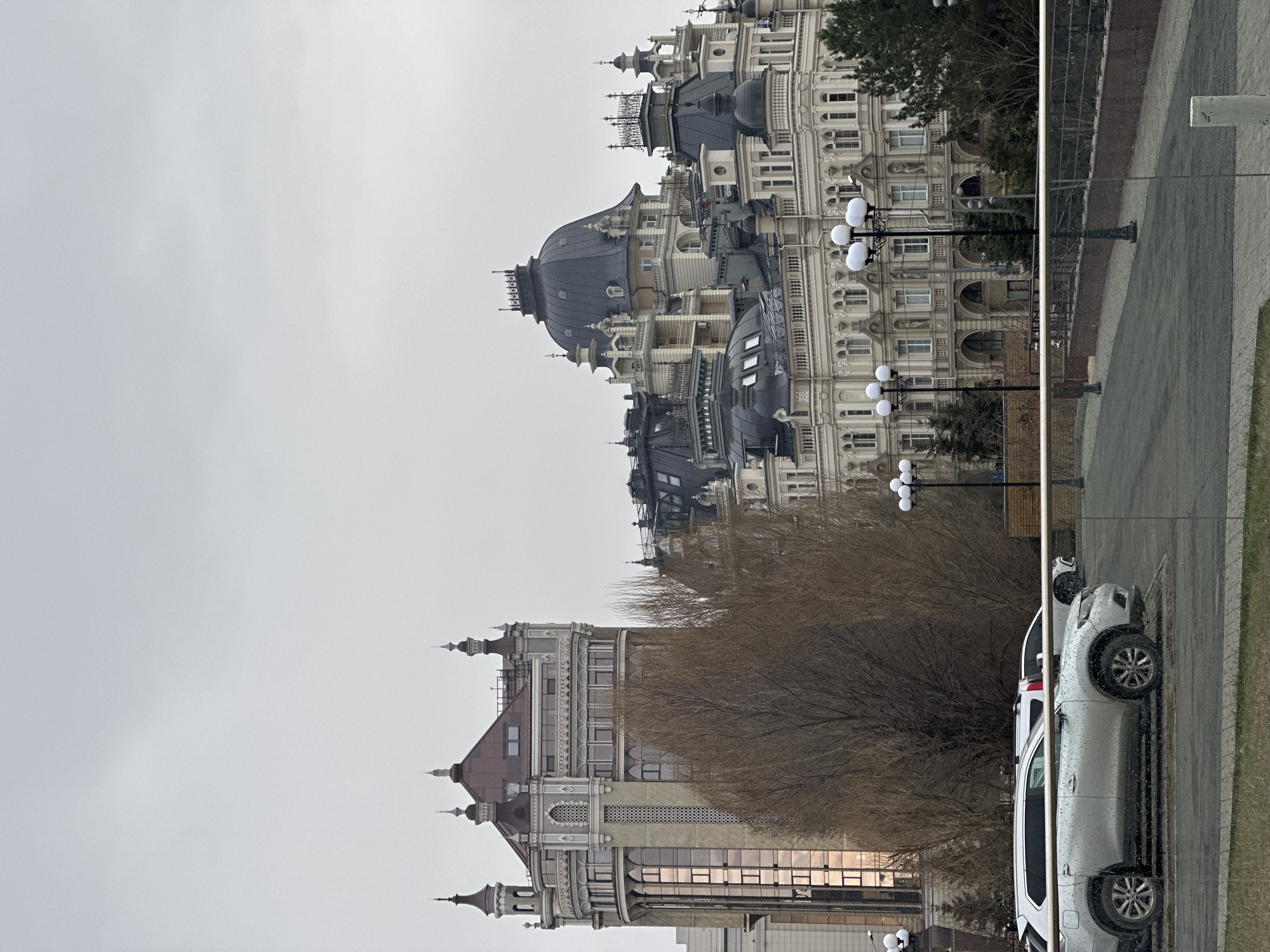 The image depicts the Palace of Farmers in Kazan, Russia. The building has a complex architectural style with multiple levels, domes, and spires. Its facade is primarily light-colored, with dark-colored domes. In the foreground, bare trees are visible. A grey paved area is present, and a silver car is parked alongside it. The sky is overcast and grey. The image appears to be taken through a window, as a vertical divider is visible, and the reflections suggest the presence of glass.