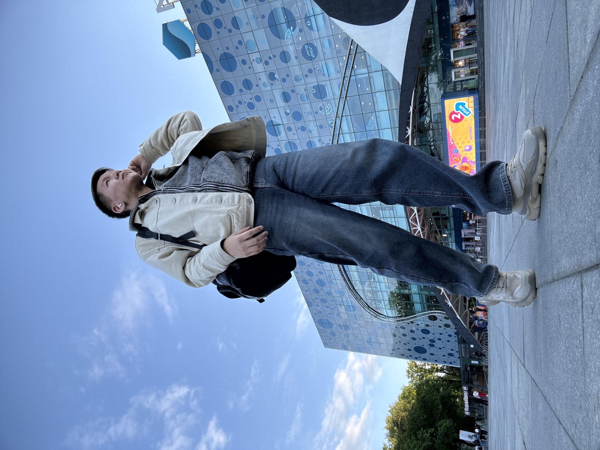 The image, taken from a low-angle perspective, depicts a young man standing on a paved surface, looking upwards towards a large, modern building under a clear blue sky with scattered white clouds. The man, positioned centrally, is dressed in a light-colored jacket over a grey hooded top, dark blue jeans, and chunky light-beige sneakers. He wears a black backpack and has a wireless earbud in his right ear, with his left hand near his face.
The prominent building in the background features a distinctive glass facade adorned with a pattern of varying blue circles and subtle aquatic-themed designs, possibly representing marine life. On a lower section of the building, a large digital screen is visible, displaying vibrant colors and Russian text. Recognizable phrases include "2 ЭТАЖ" (2nd FLOOR) and "ВЫСТАВКА" (EXHIBITION), along with the "Москвариум" (Moskvarium) logo. Green trees are partially visible in the lower right background, suggesting nearby greenery. The bright lighting indicates it is daytime. The man's pose suggests observation or contemplation of the building or sky above.
