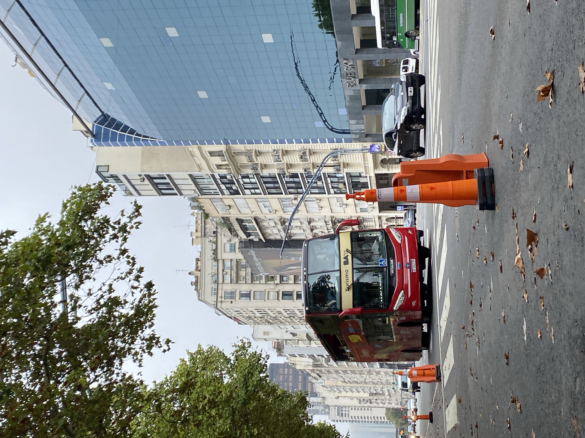 The image depicts a street scene in Buenos Aires, Argentina. A red "Turismo BA" double-decker bus is crossing a zebra crossing. Orange traffic cones are placed near the crossing. The road surface is wet with scattered leaves. A black sedan is parked on the right side of the road, and a green and white bus is partially visible behind it. Buildings are in the background, including a modern glass-faced structure. Trees are visible to the left of the frame.