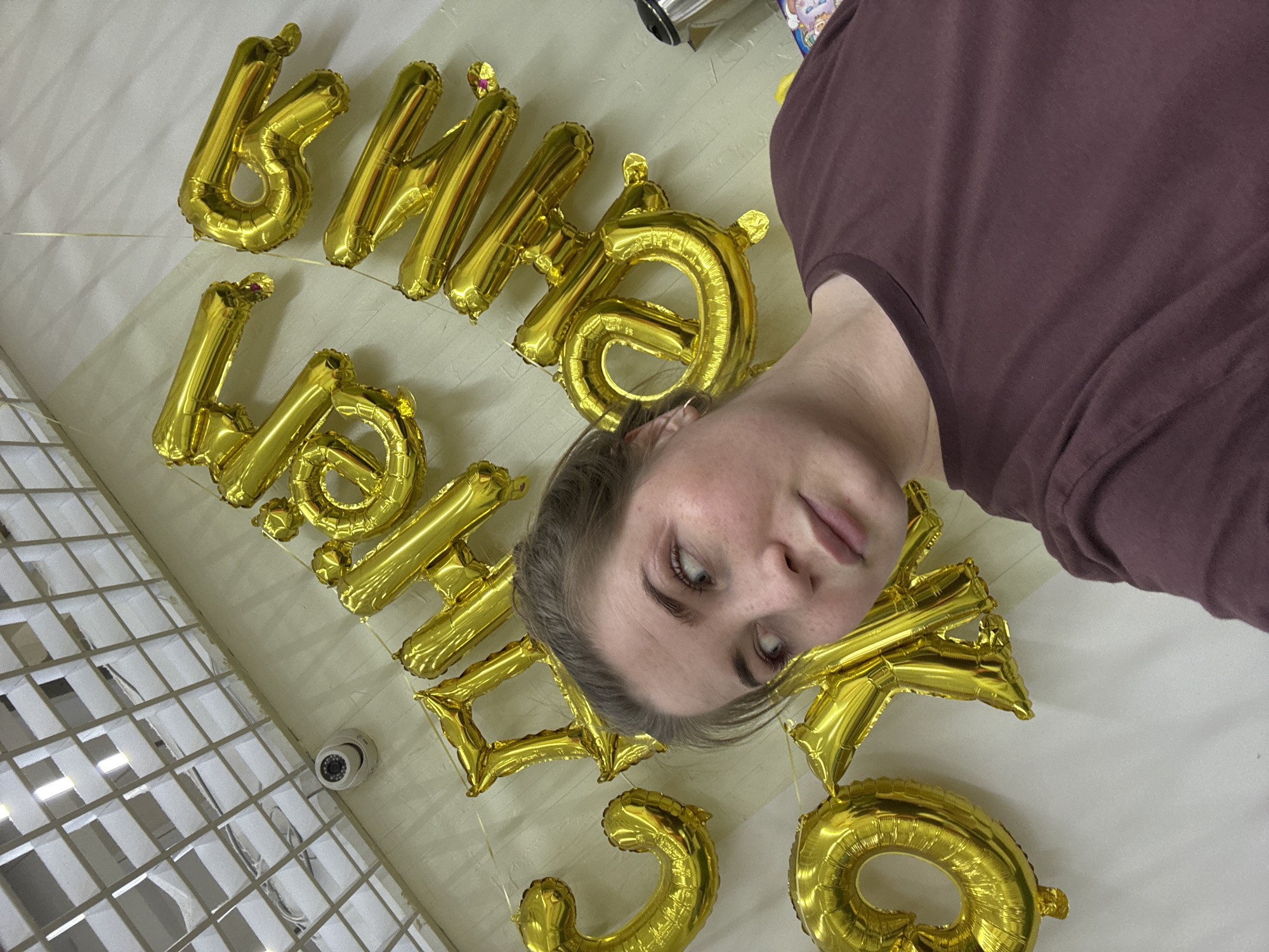 A person is lying on their back on a white surface, possibly a bed, with gold foil balloons spelling out "Happy 30" positioned around them. The person is wearing a brown top. The balloons are inflated and seem to be attached to the surface. A security camera is visible in the lower left corner of the frame, attached to a white ceiling with a grid pattern. There is no other indication of the location being in Chaikovsky, Russia.