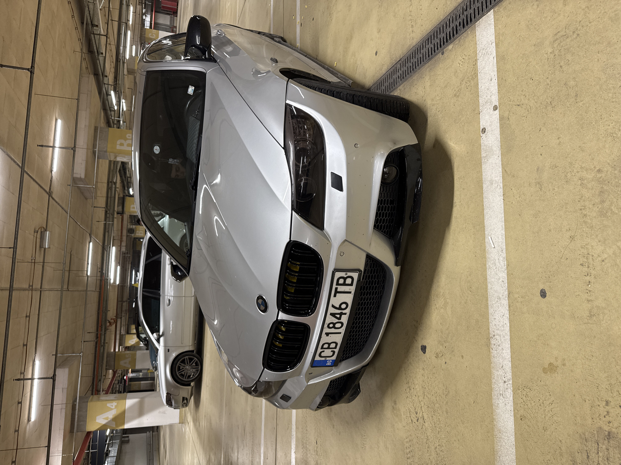 The image shows a silver BMW parked in an indoor parking garage, likely in Sofia, Bulgaria, based on the license plate "CB 1846 TB". The car is parked next to a white parking line and a drainage grate. The parking garage appears to be well-lit with overhead fluorescent lights. Another vehicle is partially visible in the background. The environment is a typical indoor parking facility.