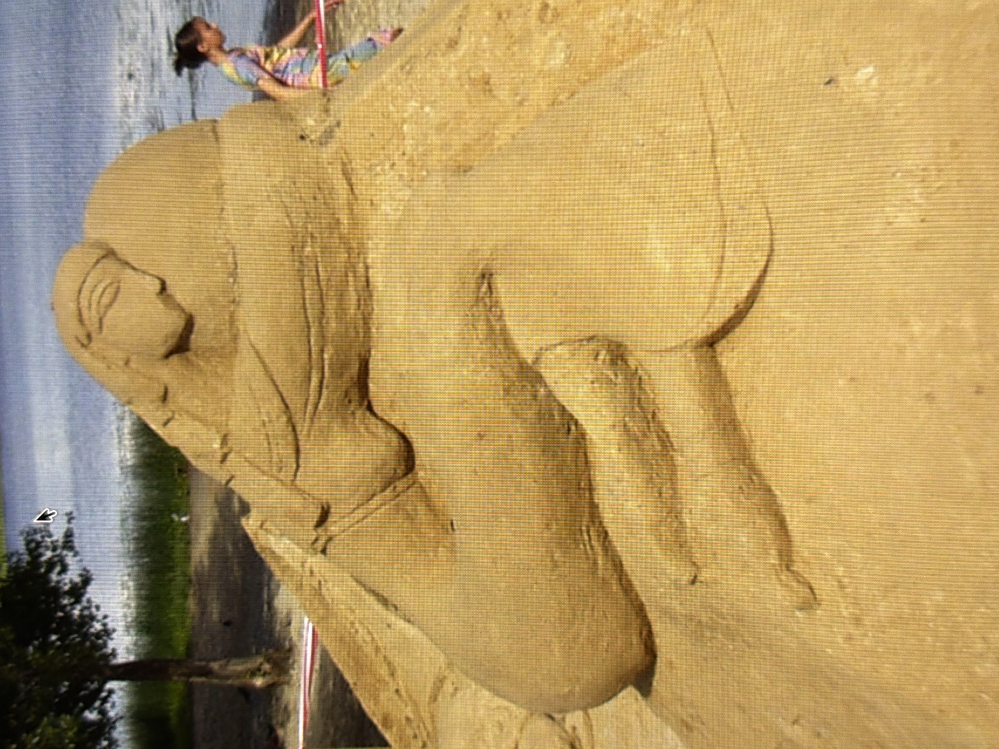 An outdoor, medium-shot, eye-level, full-color photograph shows a large sand sculpture on a sandy beach. The sculpture depicts a reclining figure with its head resting on its lap. The figure is made of sand and is light brown in color. A young girl with dark hair pulled back in a ponytail is walking behind the sculpture to the right. She is wearing a multi-colored tie-dye shirt and pants. A thin pink and red rope is stretched between two posts in front of the sand sculpture. In the background, there is a body of water, likely a river or lake, with a grassy shoreline and a tree to the left. The sky is visible in the upper part of the image. The sand sculpture is the central focus of the image. The scene appears to be a public beach or park area. The location is not specified as Kyiv, Ukraine.