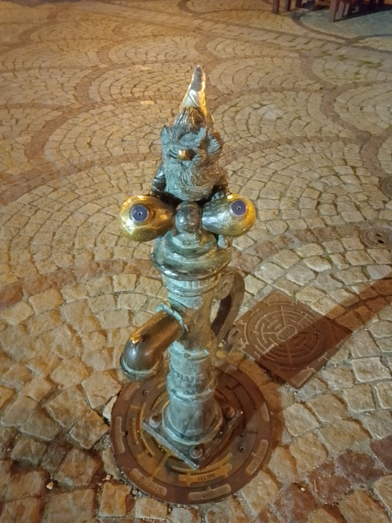 A bronze statue of a gnome is positioned on a cobblestone surface, appearing to be a functional water pump. The gnome, with a pointed hat and a beard, sits atop the pump mechanism, with its prominent golden eyes featuring blue irises. The pump itself is made of weathered bronze, showing intricate details and a spout. The cobblestone pavement is laid out in a circular pattern with alternating light and dark stones. A circular metal grate with a maze-like pattern is visible in the lower right portion of the image. The scene is lit by artificial light, casting shadows and highlighting the textures of the objects and pavement. The statue is likely located in the Old Town of Wrocław, Poland, a city known for its numerous gnome figurines that are popular tourist attractions.